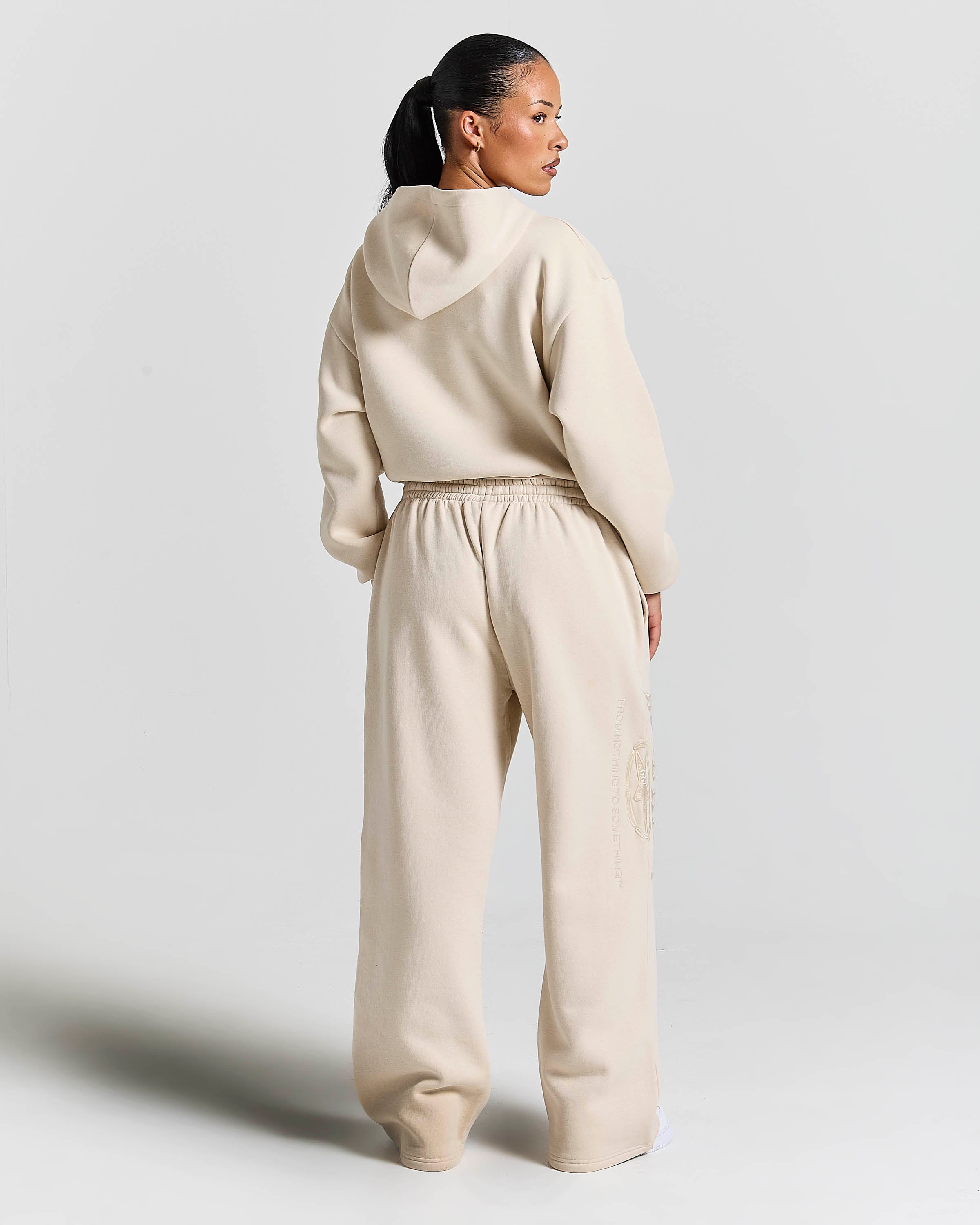 A model shown from the back wearing a matching beige cropped hoodie and wide-leg sweatpants set against a plain light gray studio background.