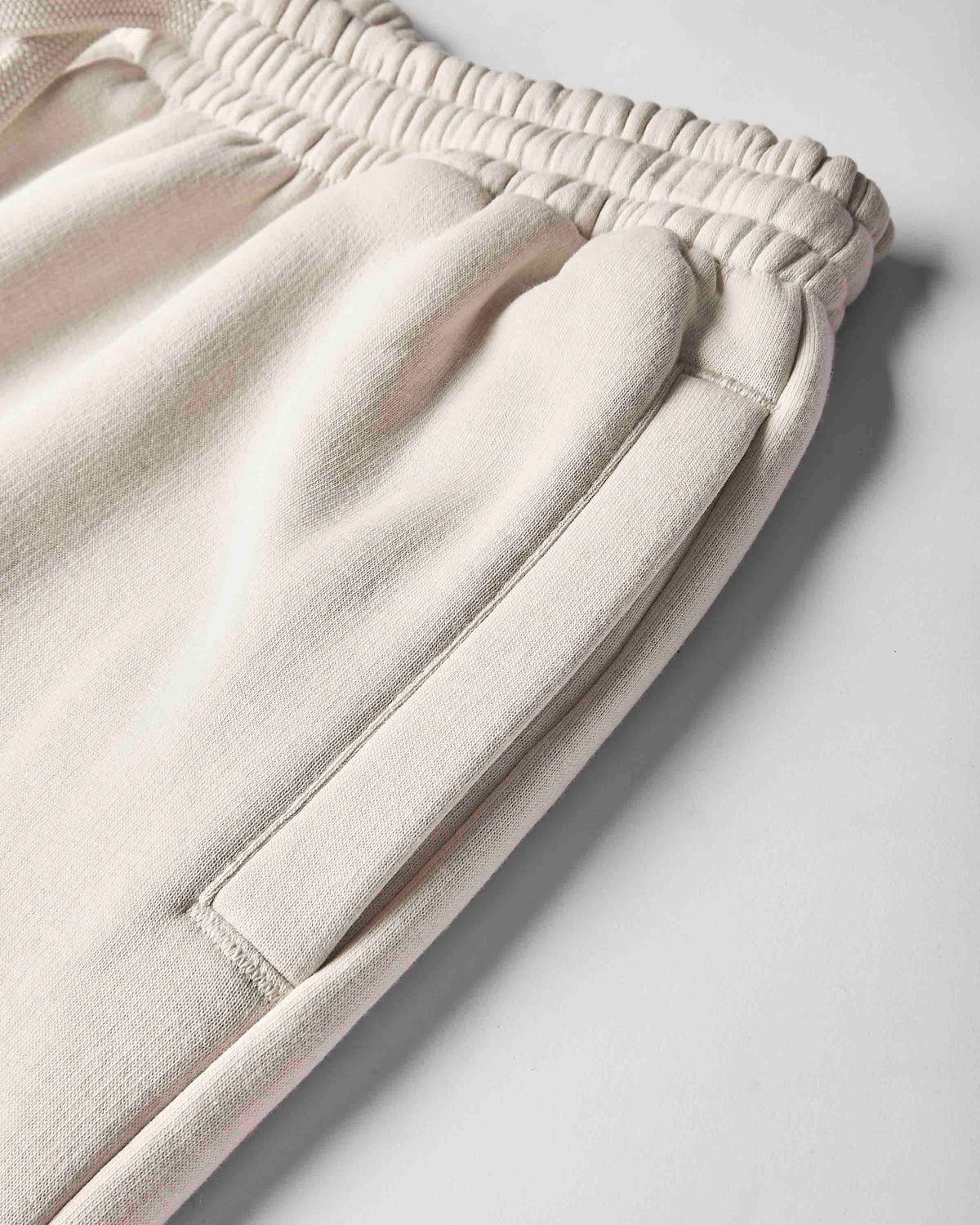 Close-up of a pair of light cream or off-white sweatpants showing the elastic waistband, side pocket seam, and soft fabric texture on a light gray background.