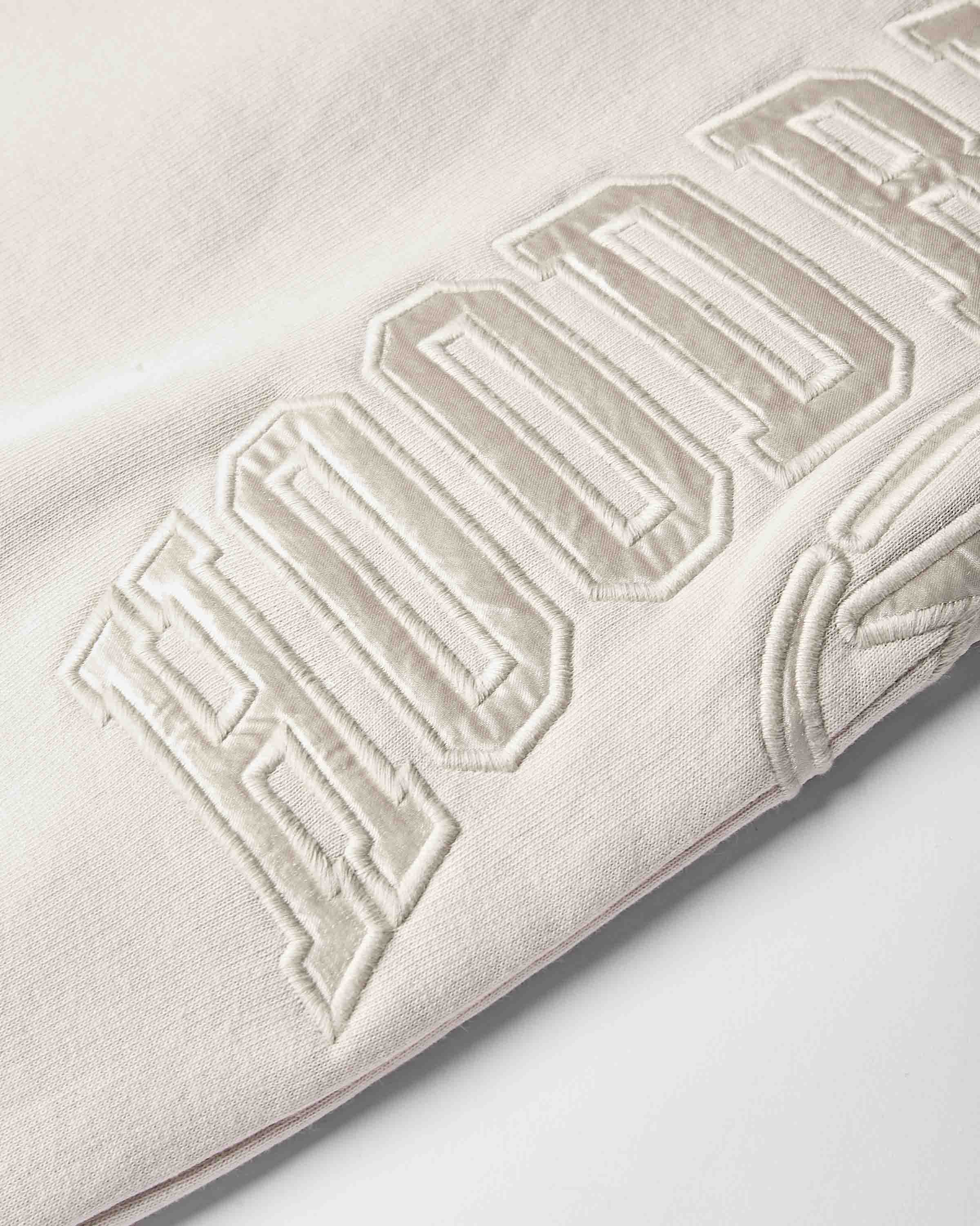 Close-up of a light cream or off-white fabric with large tonal embroidered lettering, showing the texture of the knit material and the raised embroidery.