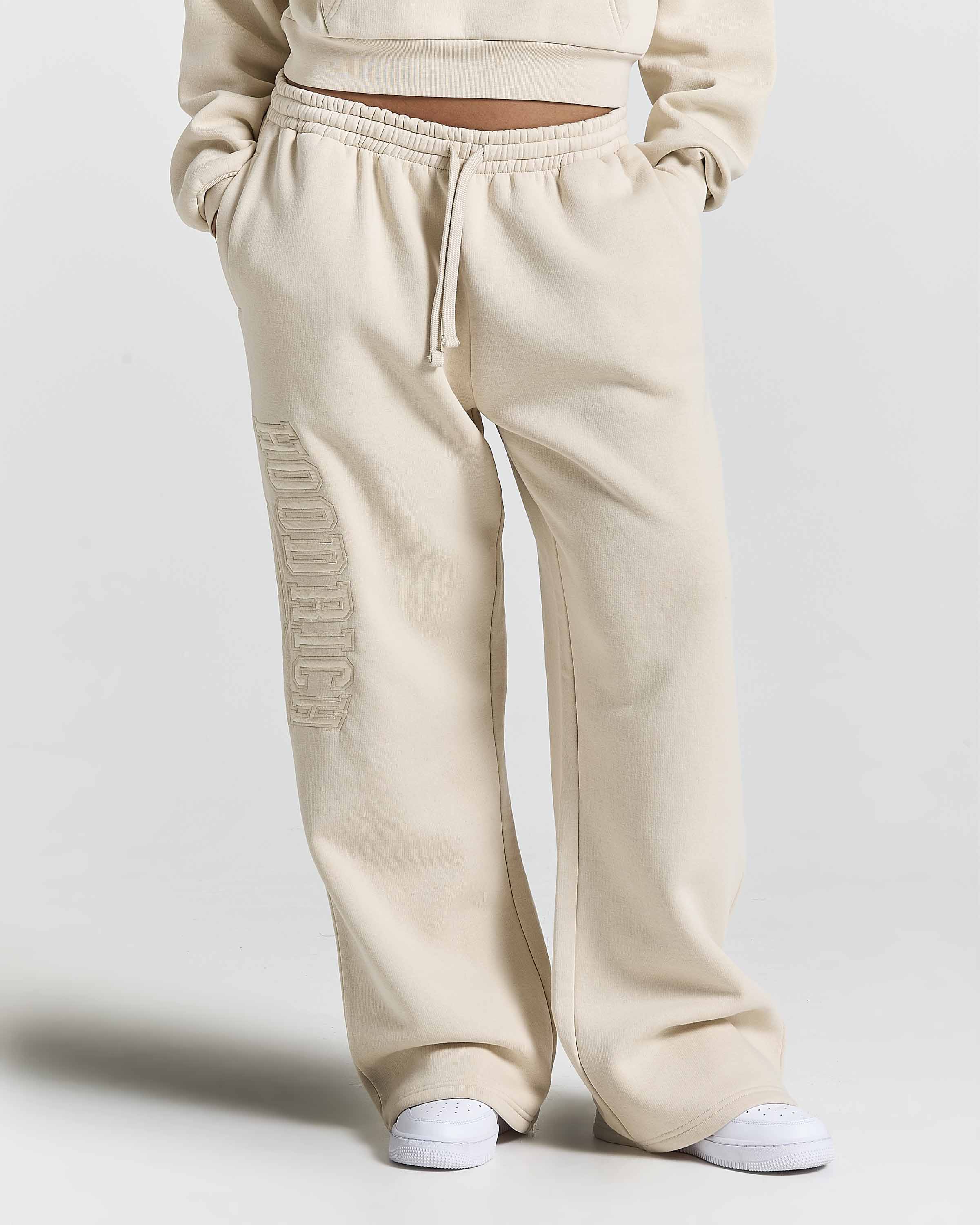 A person wearing cream-colored wide-leg sweatpants with a vertical tonal 'HOODRICH' logo on the left leg, paired with white sneakers against a light gray studio background.