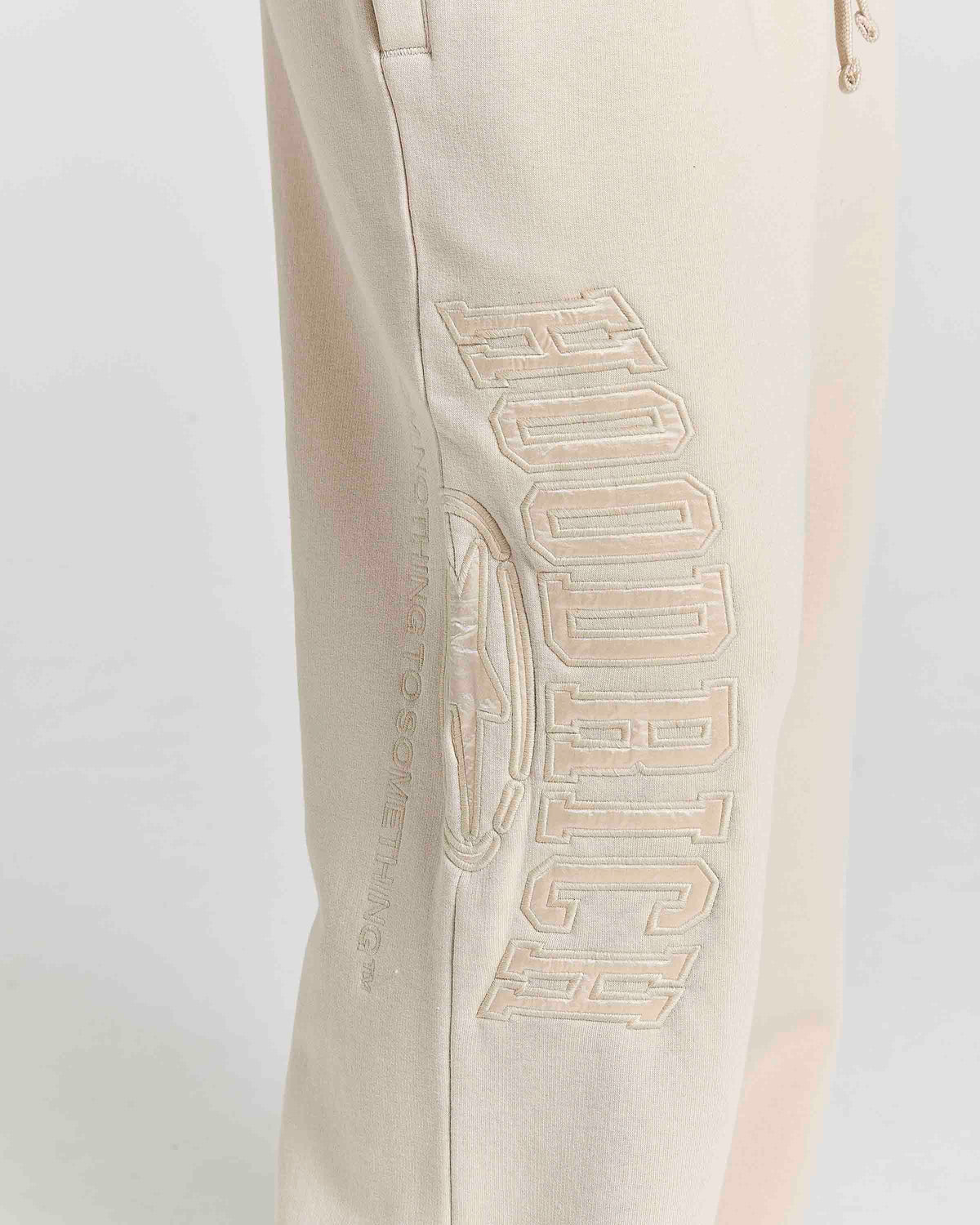 Close-up of a light beige sweatpants leg featuring a large, tonal embroidered graphic of the word 'HOODRICH' with a circular logo emblem on the side.