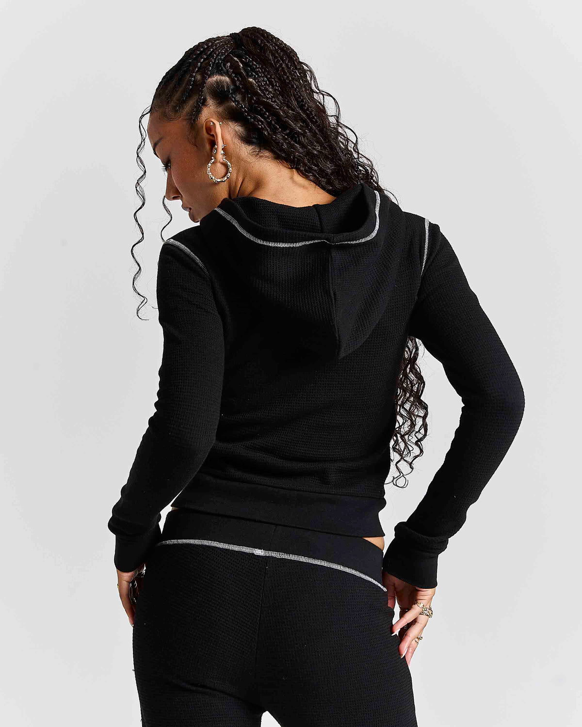 A woman shown from the back wearing a black ribbed long-sleeve hoodie with thin silver contrast piping along the hood and waist, paired with black leggings, against a light gray studio background.