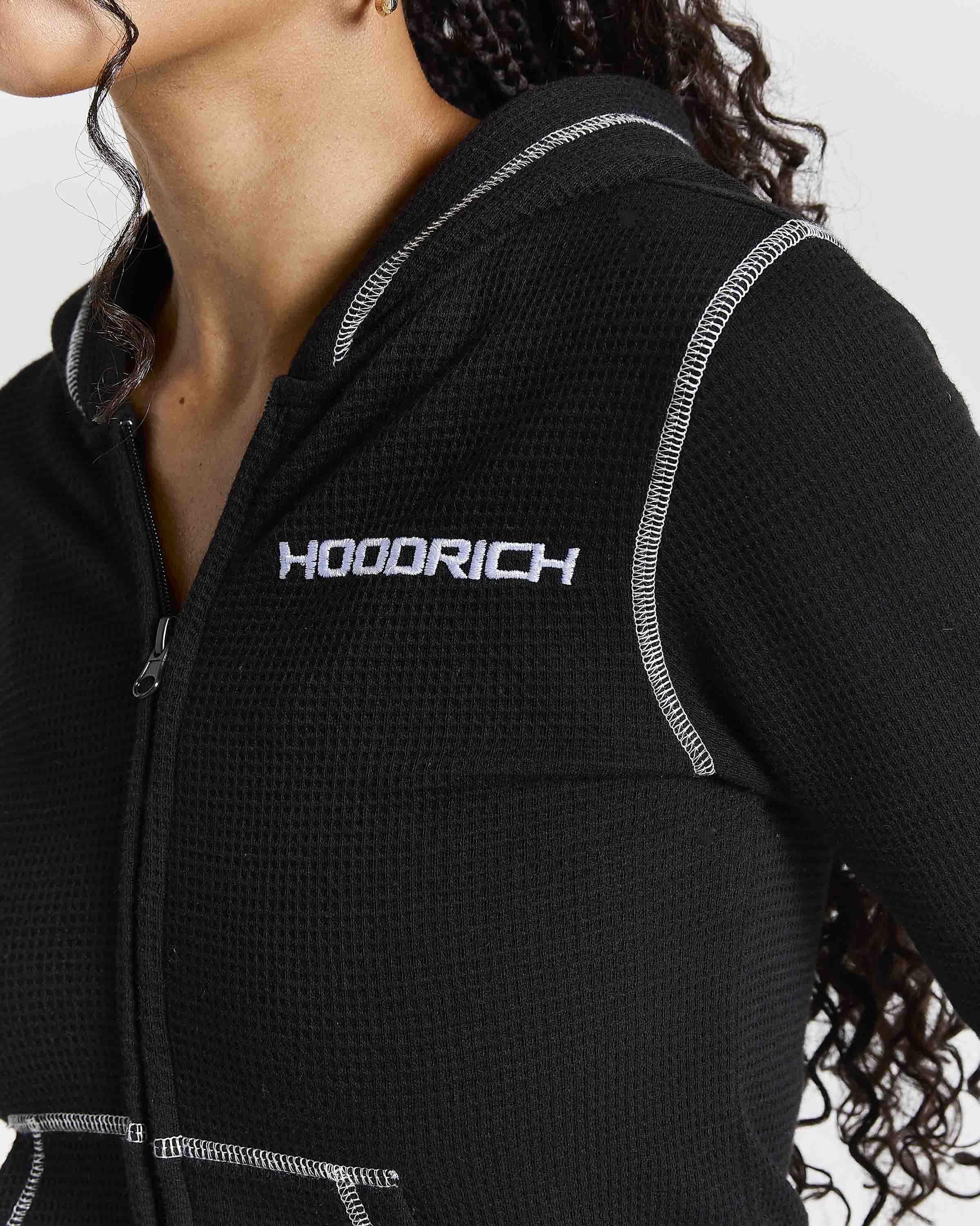 Close-up of a person wearing a black waffle-knit zip-up hoodie with white contrast stitching and a white HOODRICH logo on the chest.