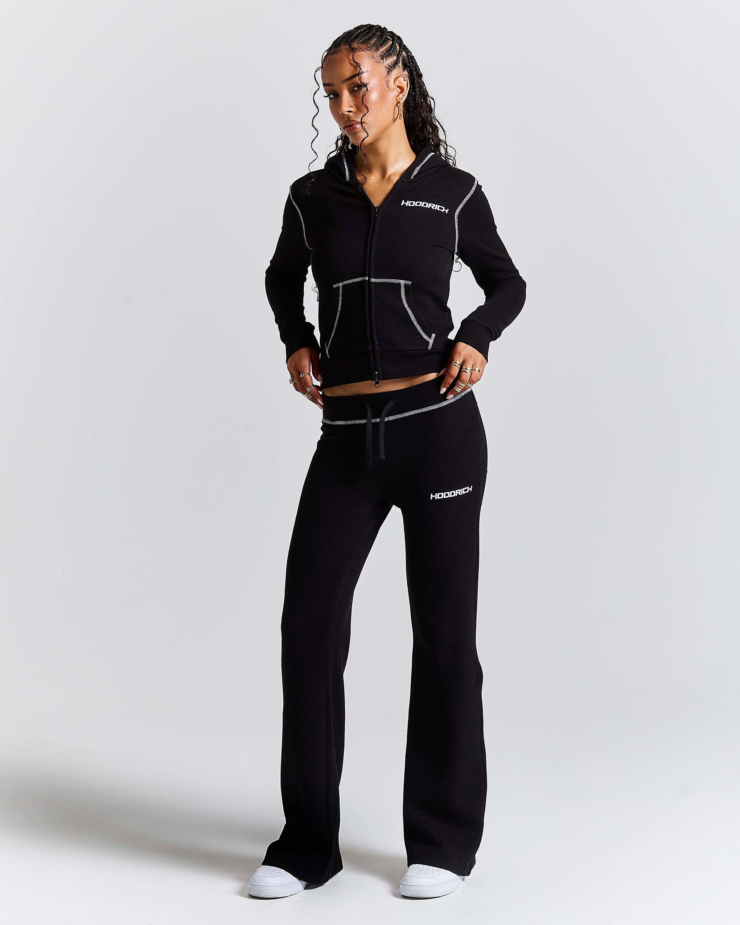 A woman modeling a black athletic tracksuit with white contrast stitching and white logo text on the chest and thigh, photographed against a light gray studio background.