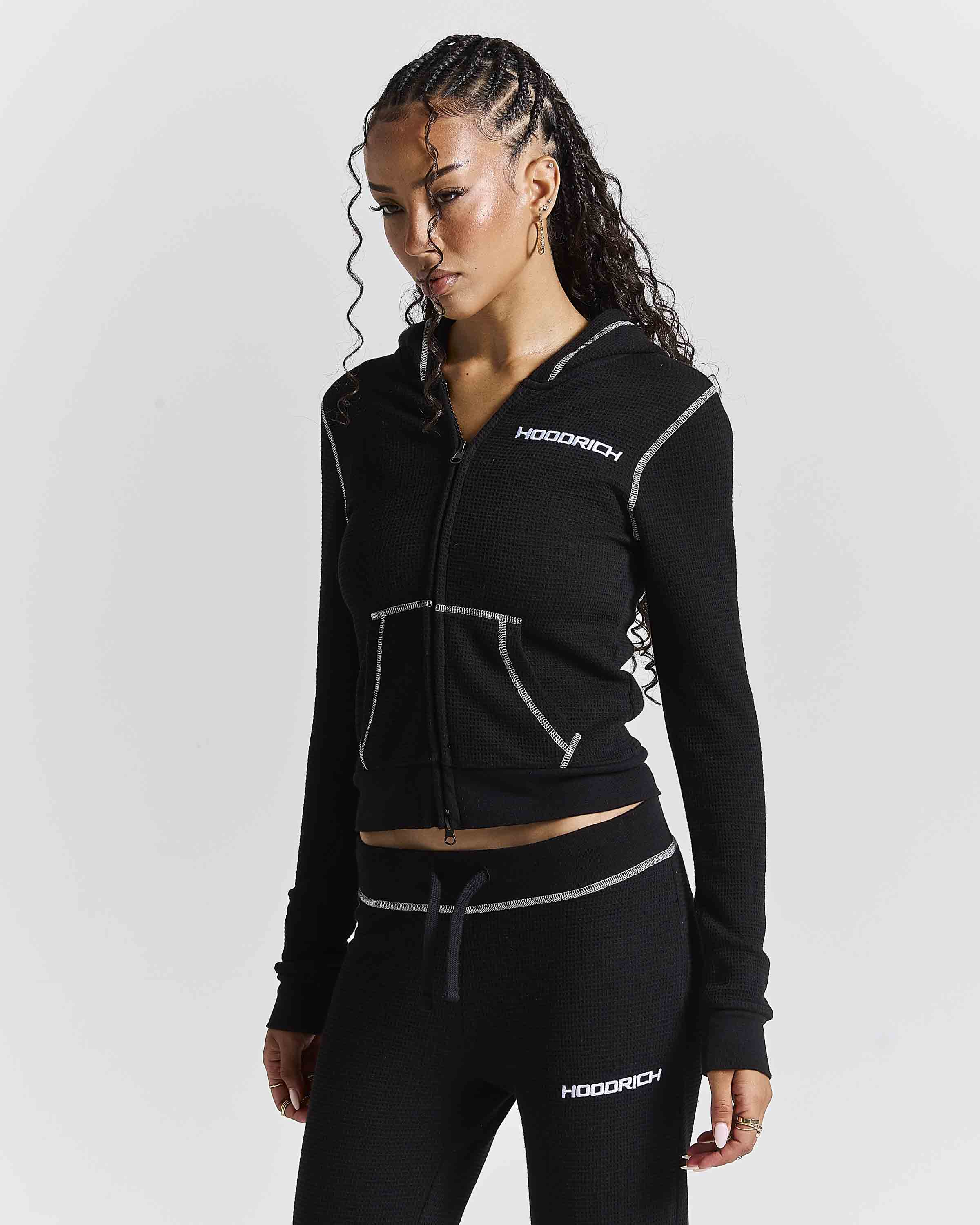 A woman modeling a black zip-up hoodie and matching black joggers with white contrast stitching and white HODGICH logos, photographed against a light gray studio background.