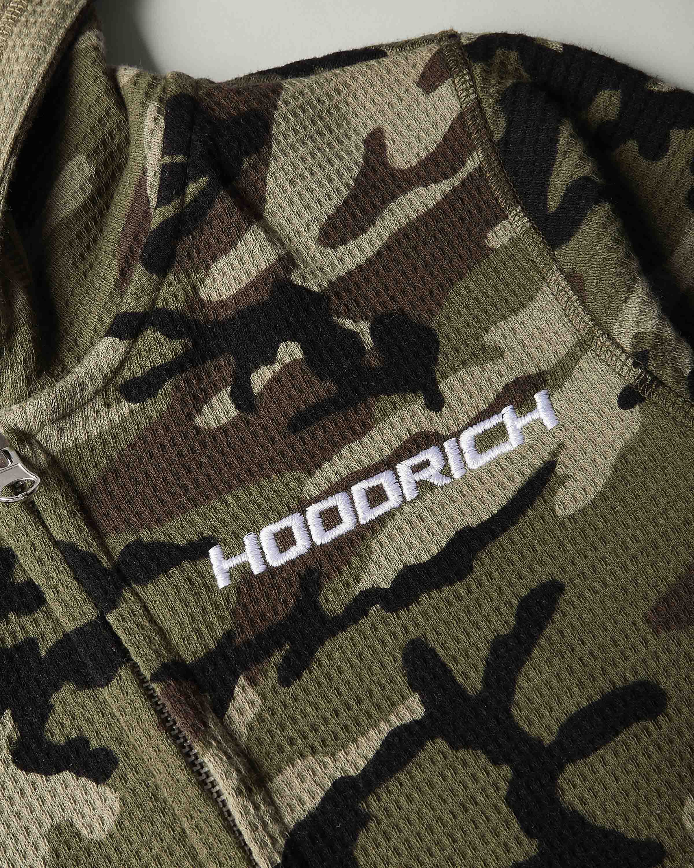 Close-up of a camouflage mesh zip jacket with a white embroidered 'HOODRICH' logo on the chest.