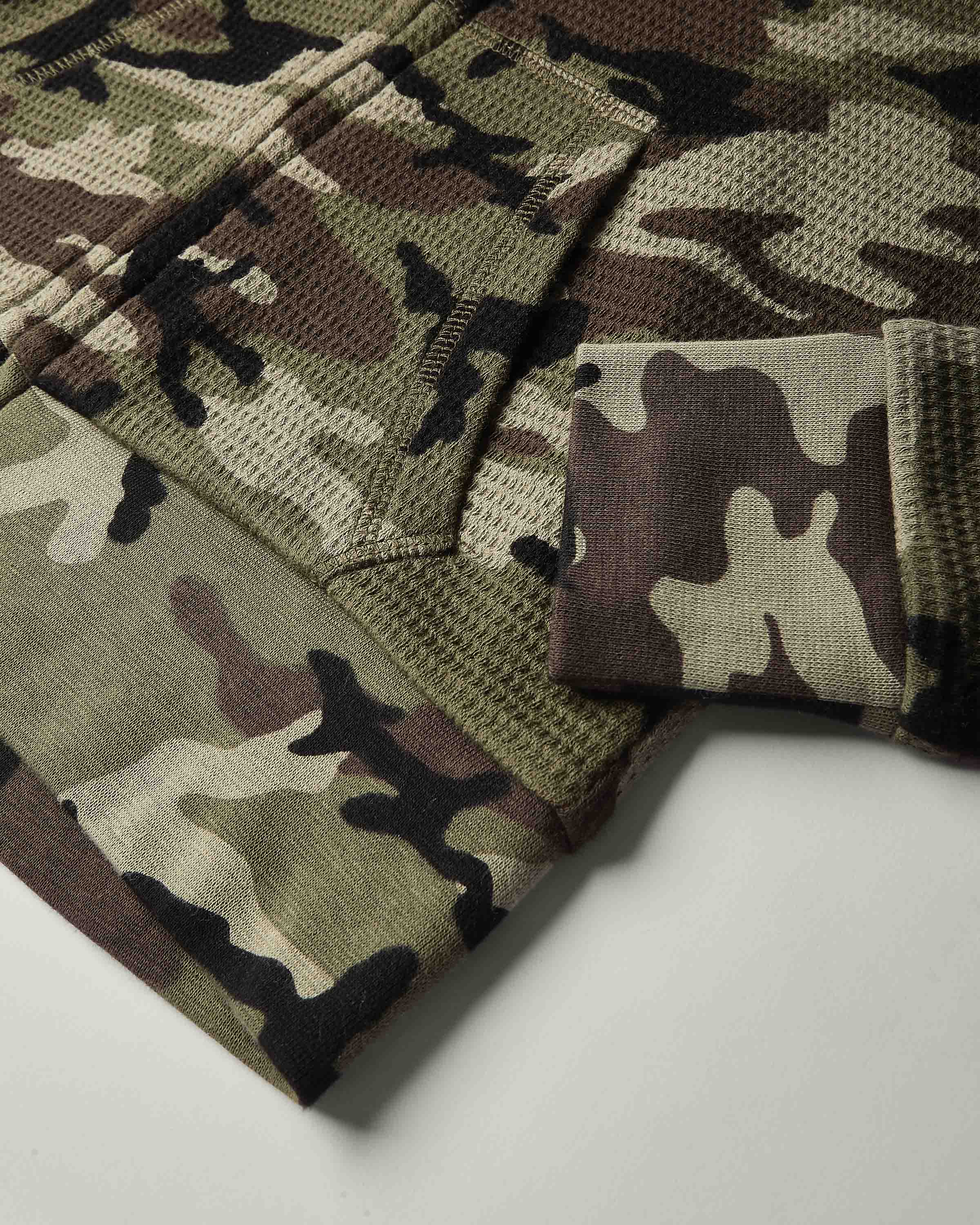Close-up of a camouflage knit fabric garment, showing a waffle-knit texture with a pocket and folded cuff in shades of olive green, brown, beige, and black.