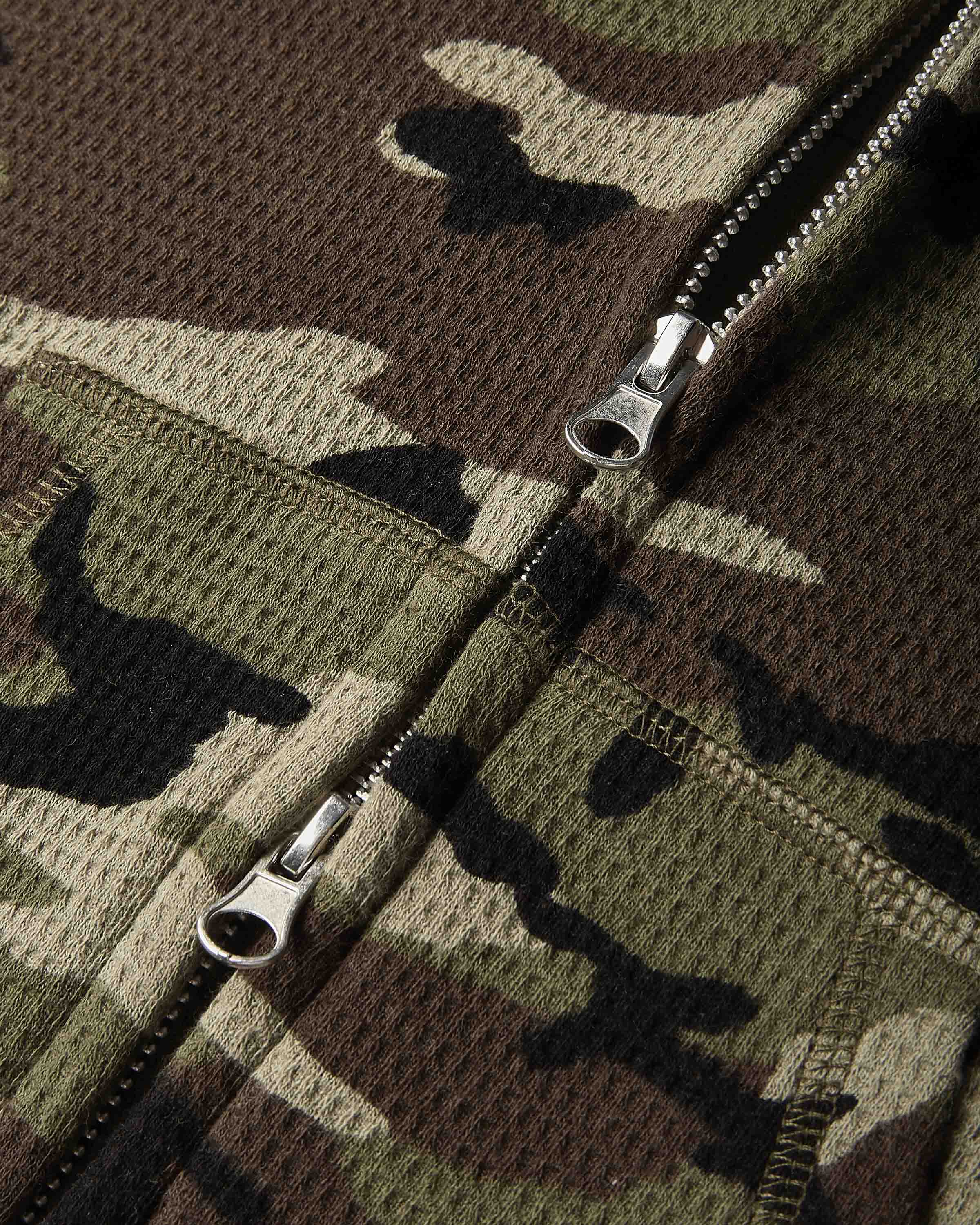 Close-up of a camouflage zippered garment, showing a textured waffle-knit fabric in green, brown, black, and beige tones with silver metal zipper pulls.
