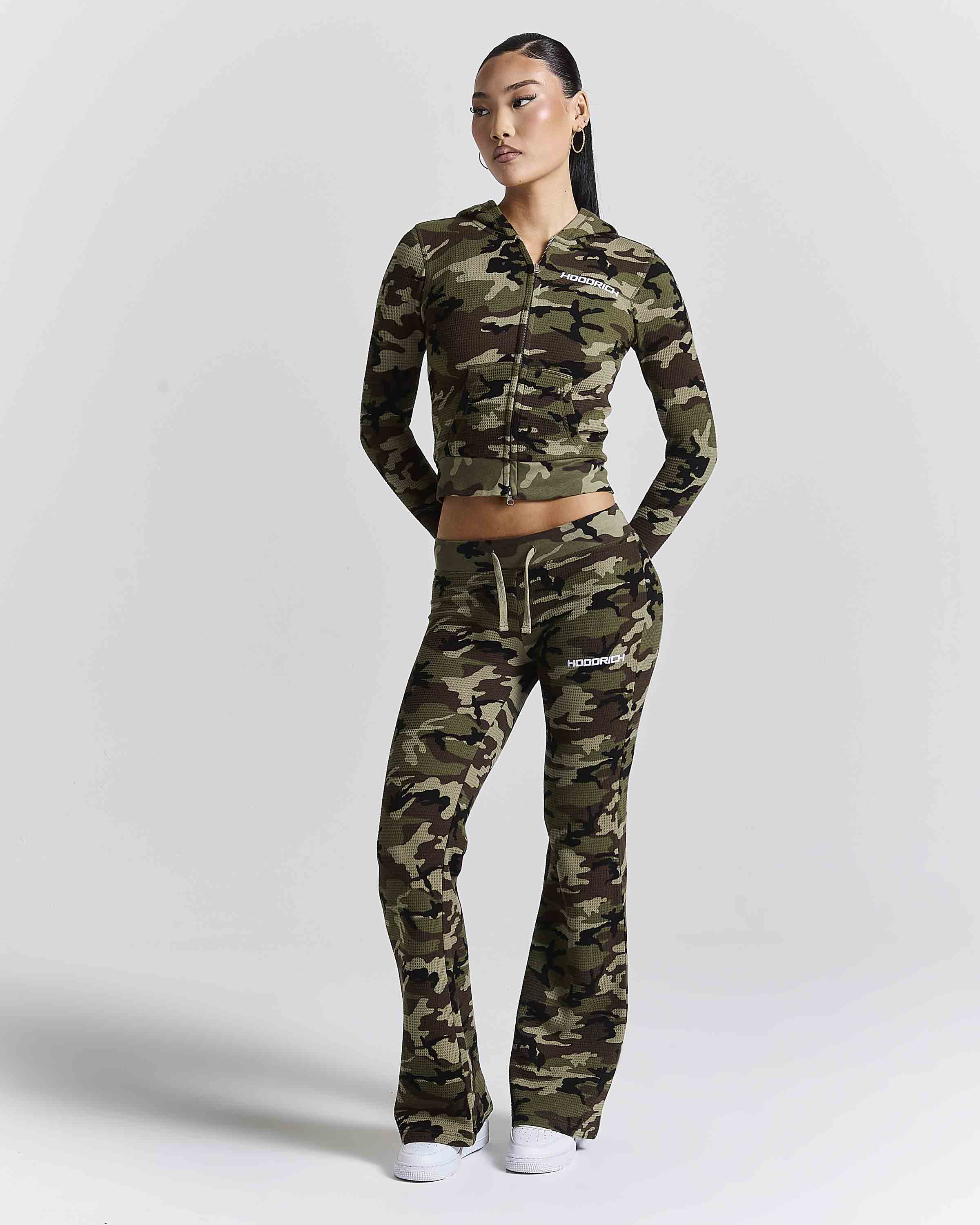 A model wearing a matching camouflage zip-up hoodie and flared sweatpants set, standing against a light gray studio background.