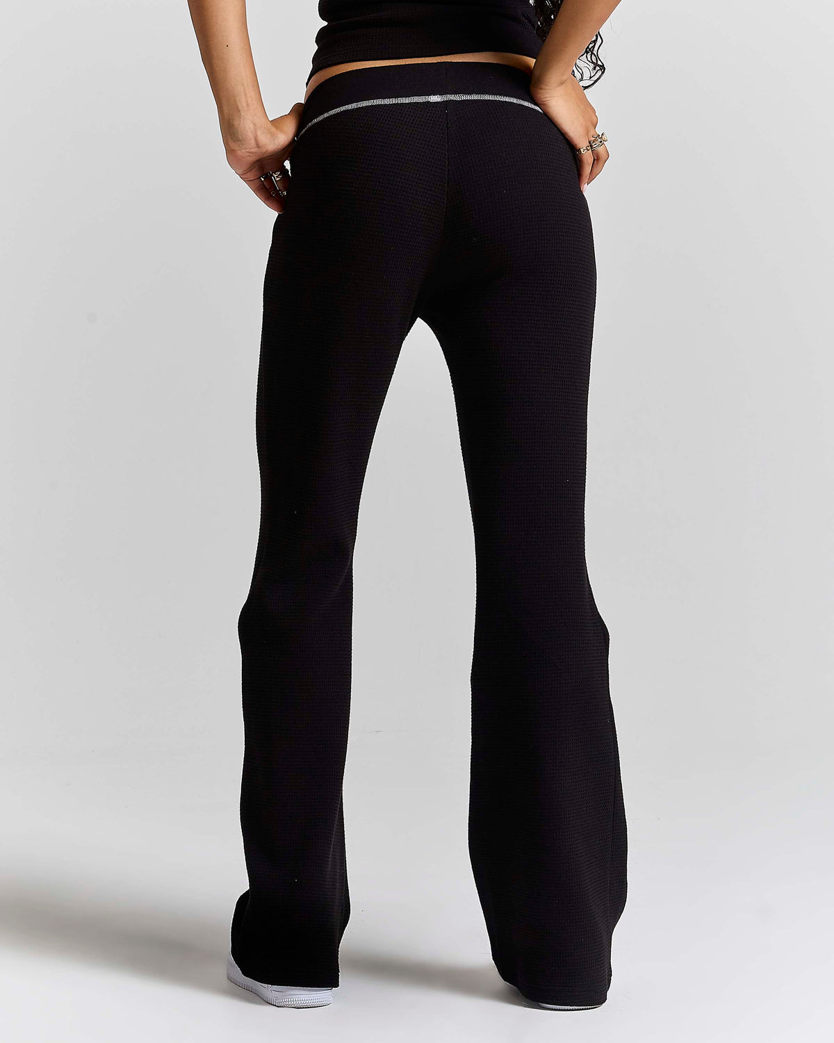 Rear view of a model wearing black flared pants with a thin silver metallic trim detail at the back waistband, set against a plain light gray studio background.