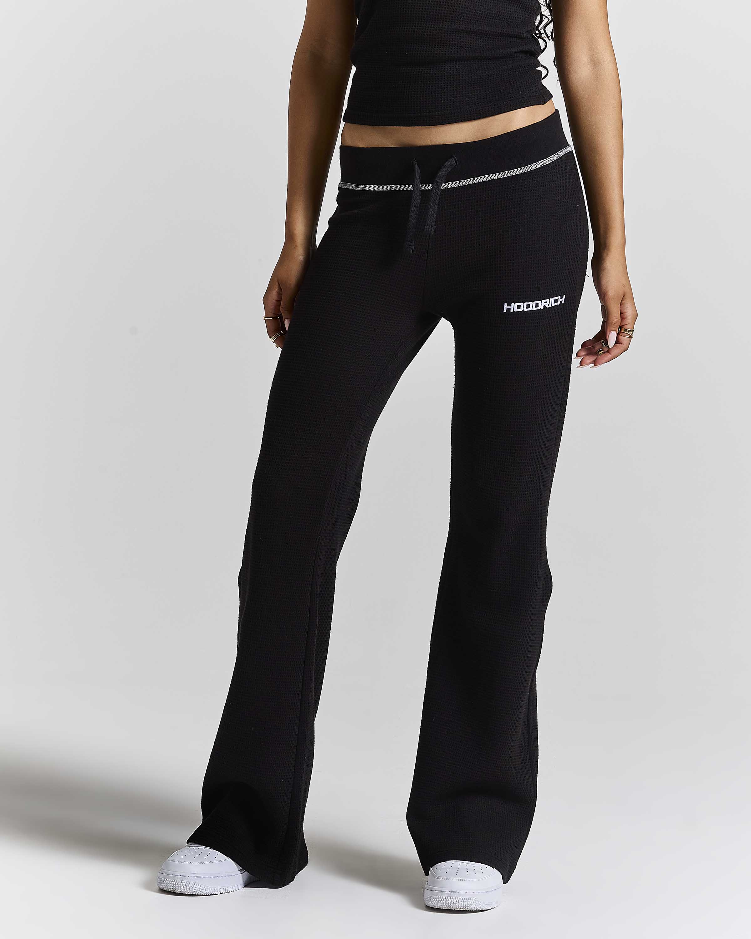 A model wearing black wide-leg sweatpants with a white drawstring and a white 'HOODRICH' logo on the left thigh, paired with a black crop top and white sneakers, set against a plain light gray studio background.