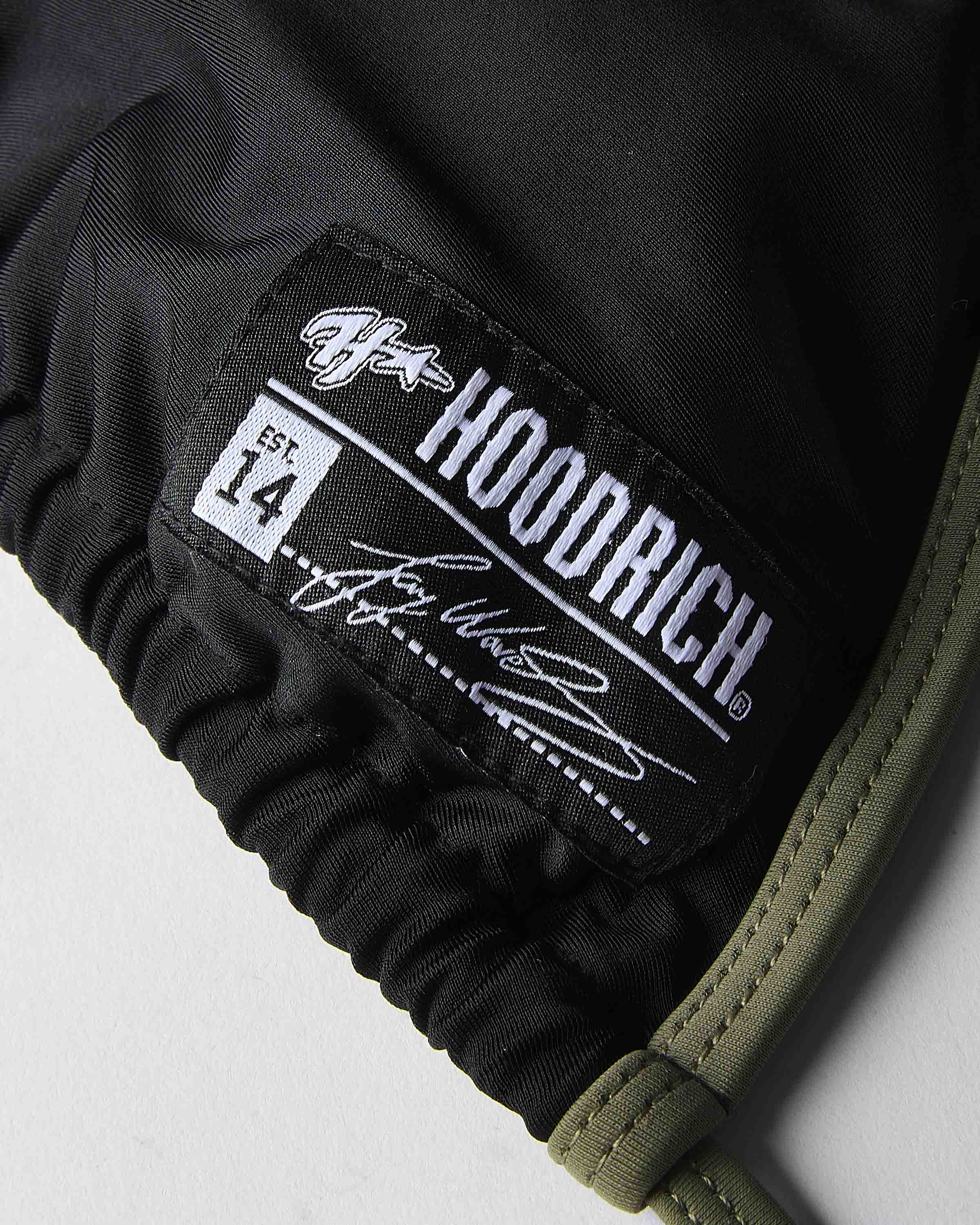 Close-up of a black hooded garment with a sewn-on brand patch reading Hoodrich , plus an olive green ribbed trim along the edge.