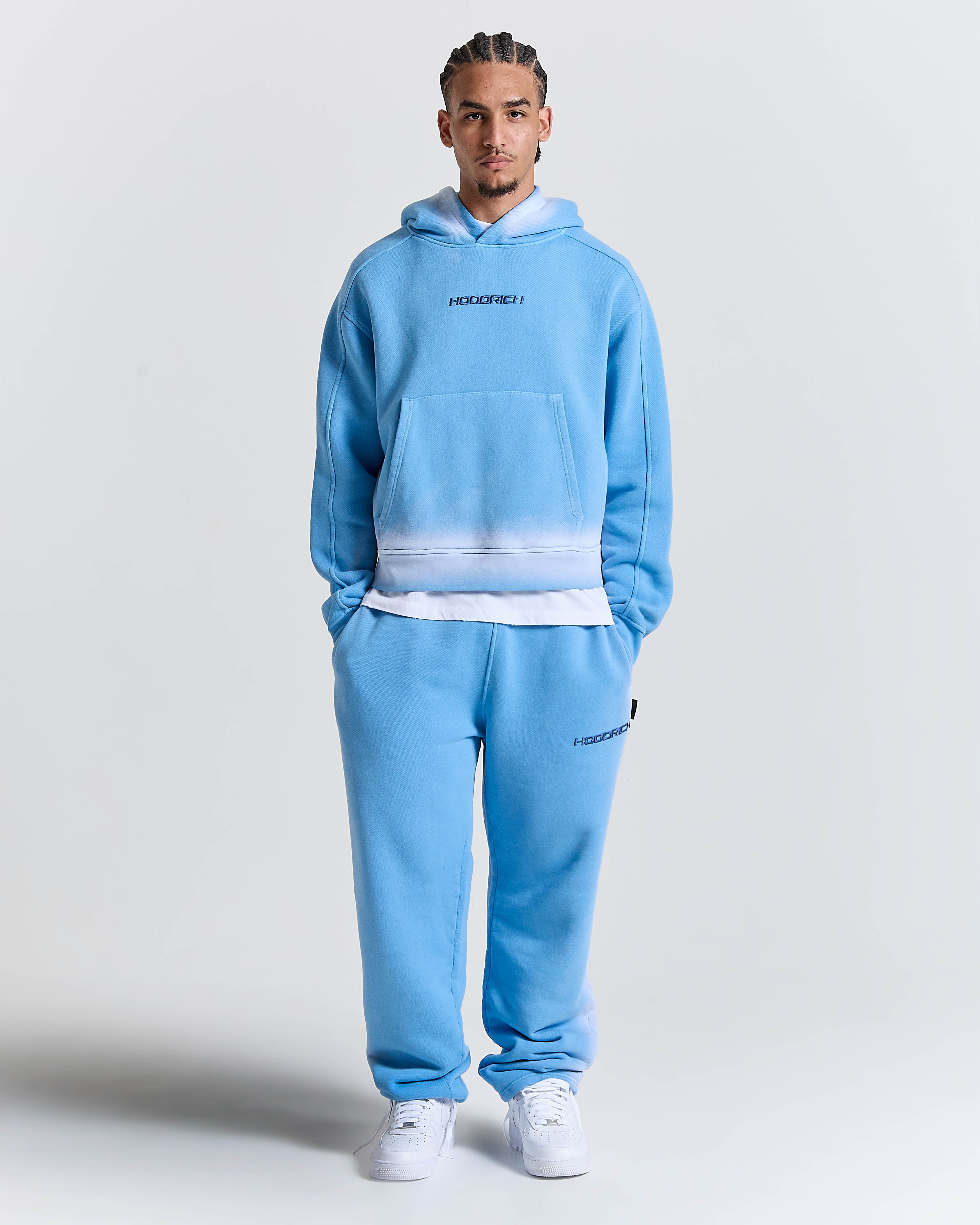 Model wearing oversized blue joggers featuring a black Hoodrich logo embroidery on the thigh, white socks, and white trainers.