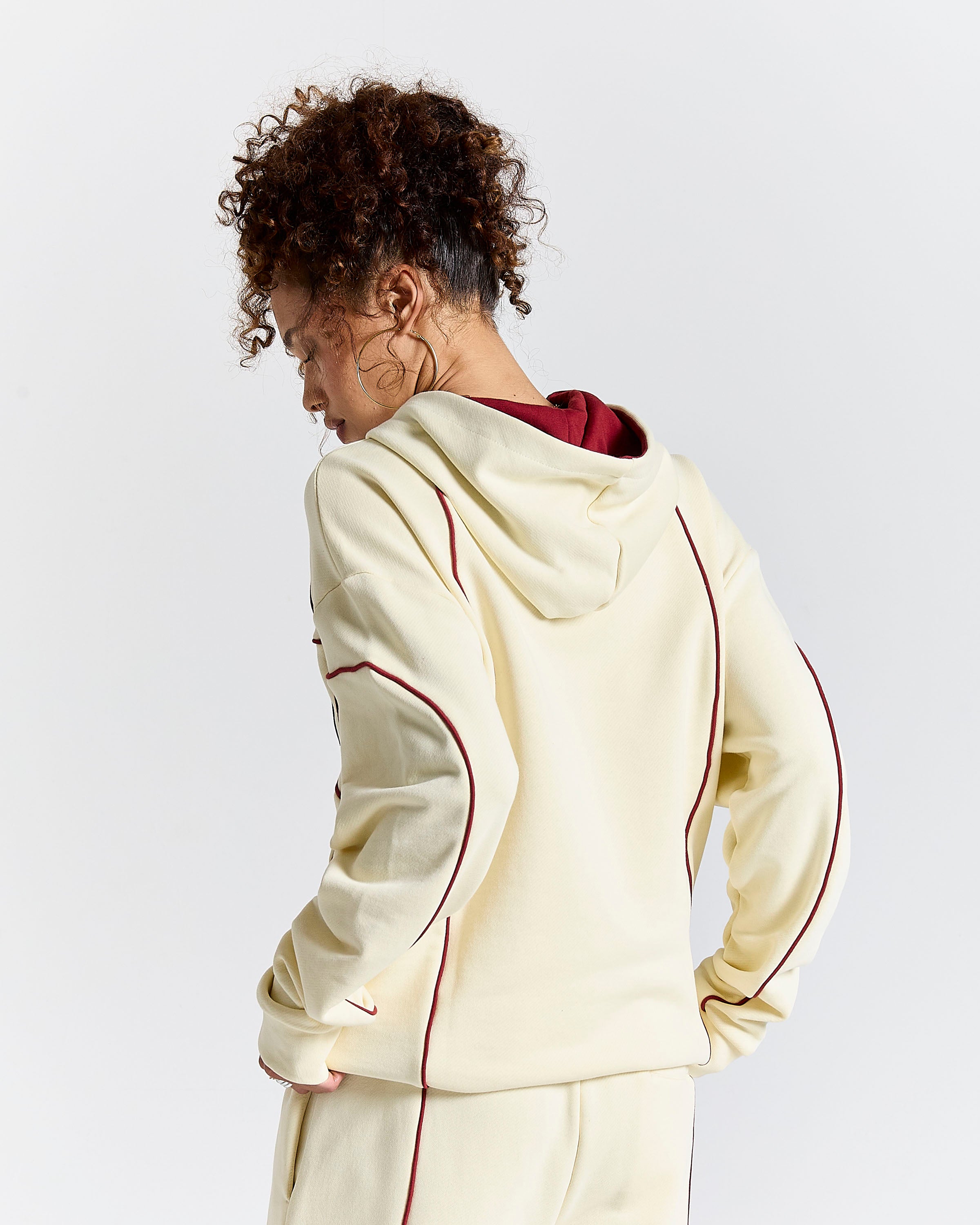 A model shown from the back wearing a cream-colored Hoodrich hoodie with burgundy piping and lining, paired with matching sweatpants, against a light gray studio background.
