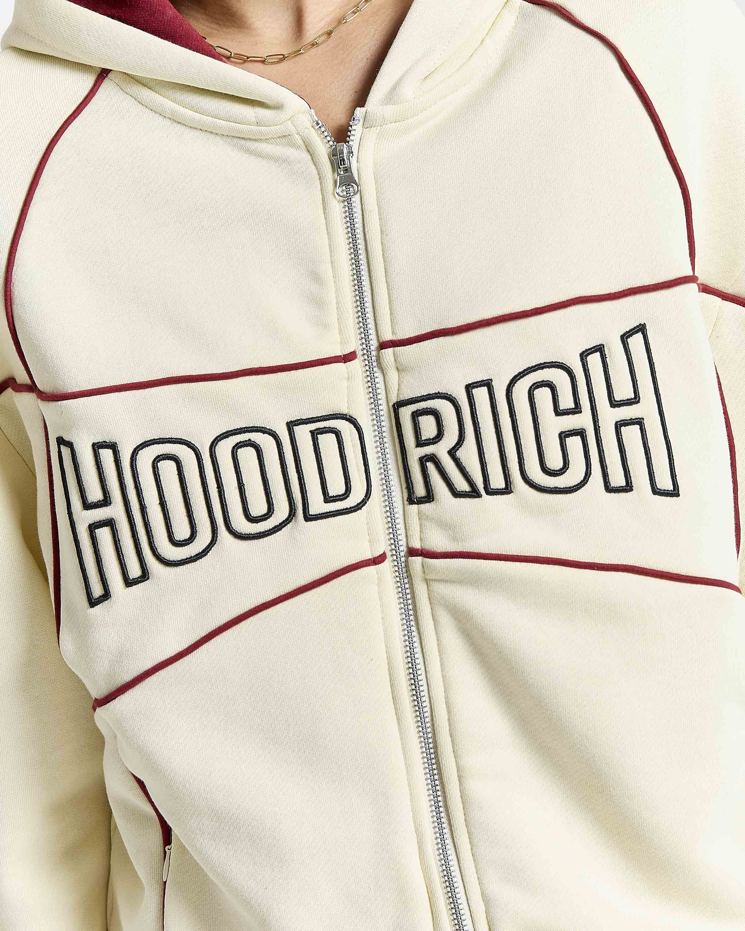 Close-up of a cream zip-up hoodie with 'HOOD RICH' embroidered across the chest in black outlined letters, accented by dark red piping lines.