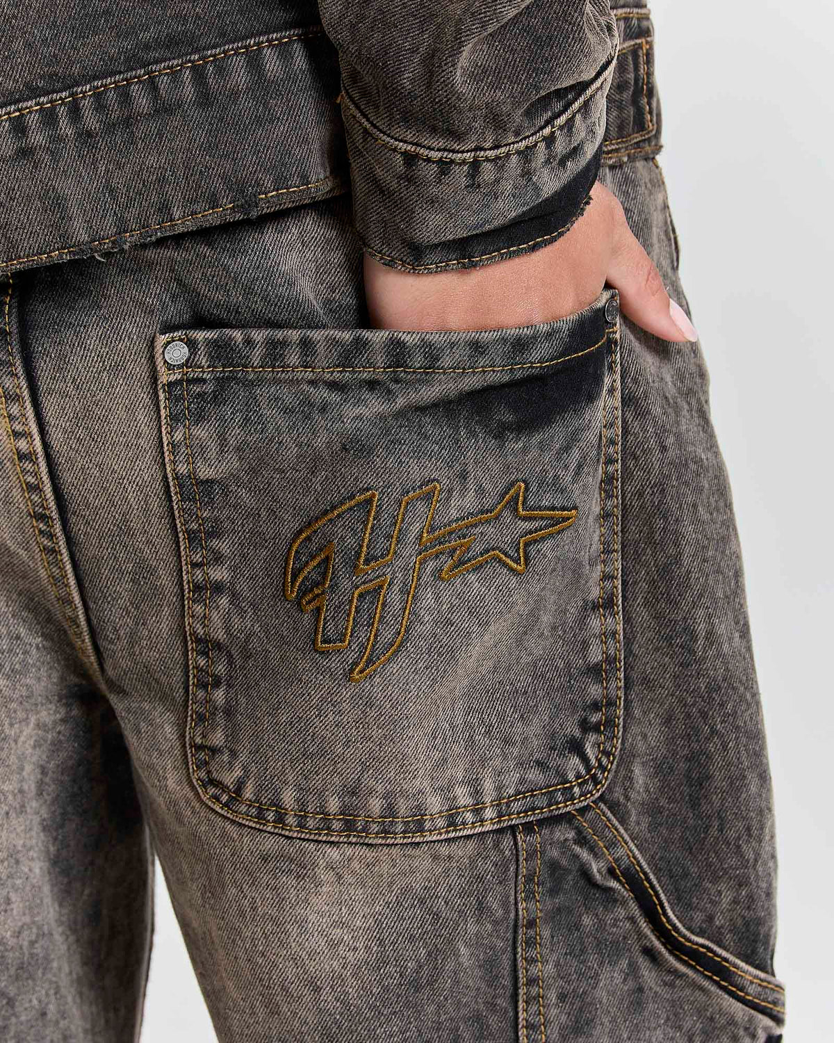 Close-up of a person wearing washed charcoal gray denim jeans with a gold embroidered Hoodrich logo on the back pocket, showing a hand in the pocket.