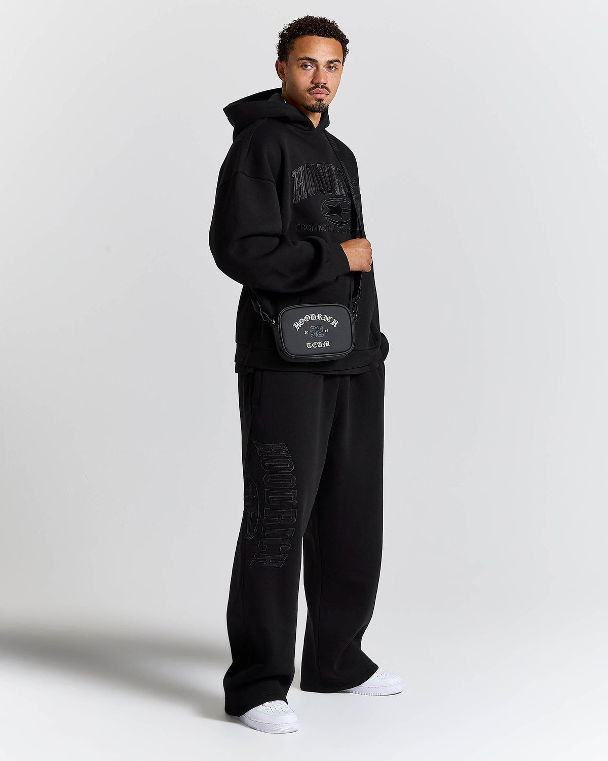A man modeling a black streetwear outfit from the brand HOODRICH, featuring a black hoodie with tonal graphic lettering, matching black sweatpants with tonal graphic details, and a black crossbody bag with white branding.
