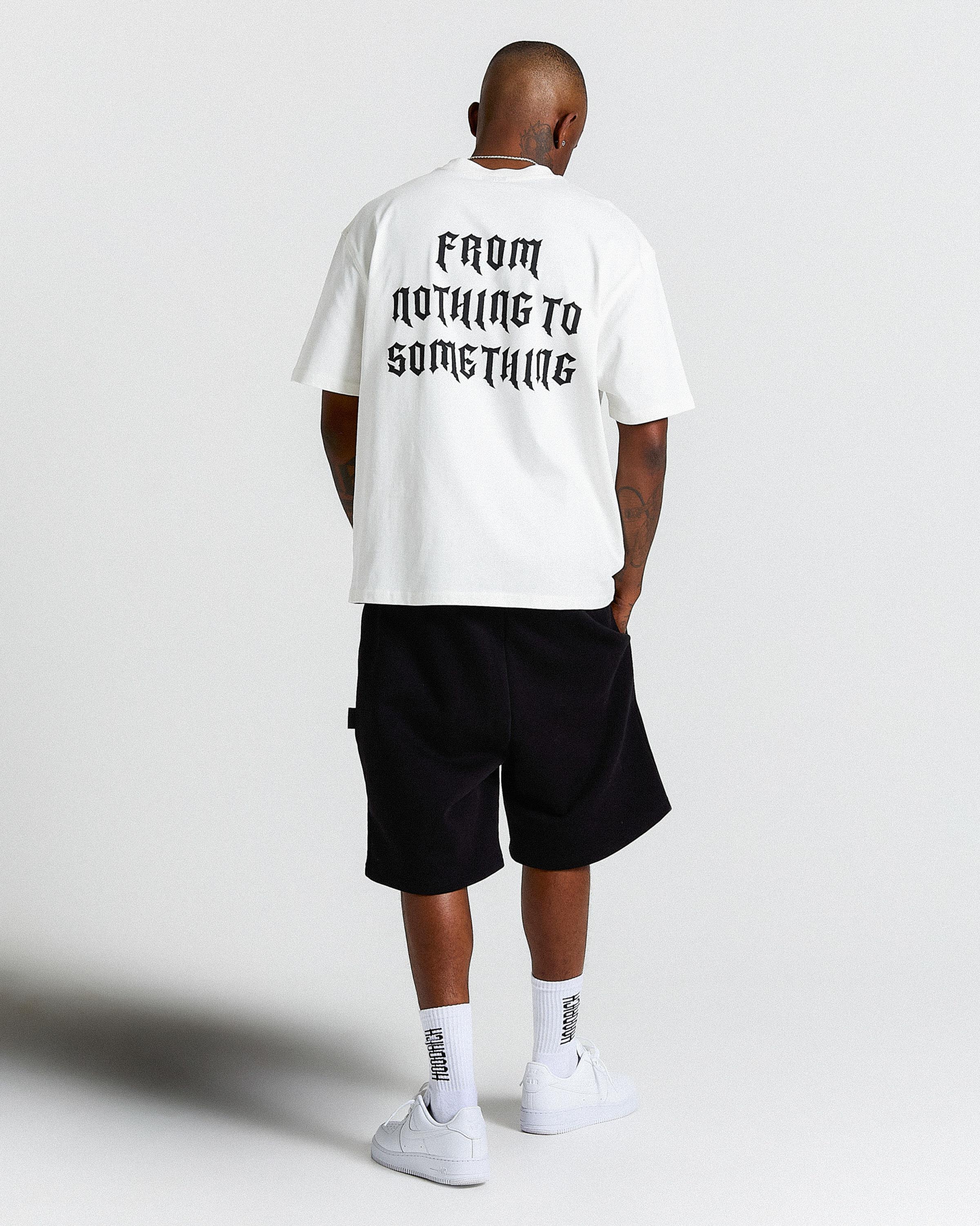 Person wearing a white t-shirt with text on the back, black shorts, and white sneakers on a light gray background