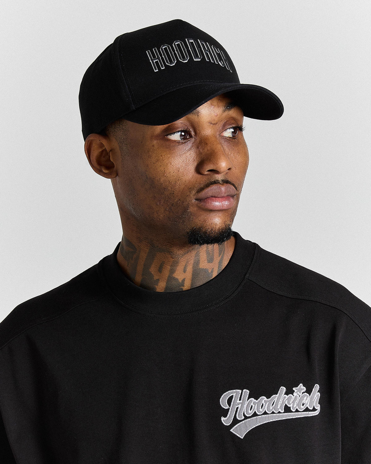 Man wearing a black cap and black sweatshirt with 'Hoodrich' branding on a gray background