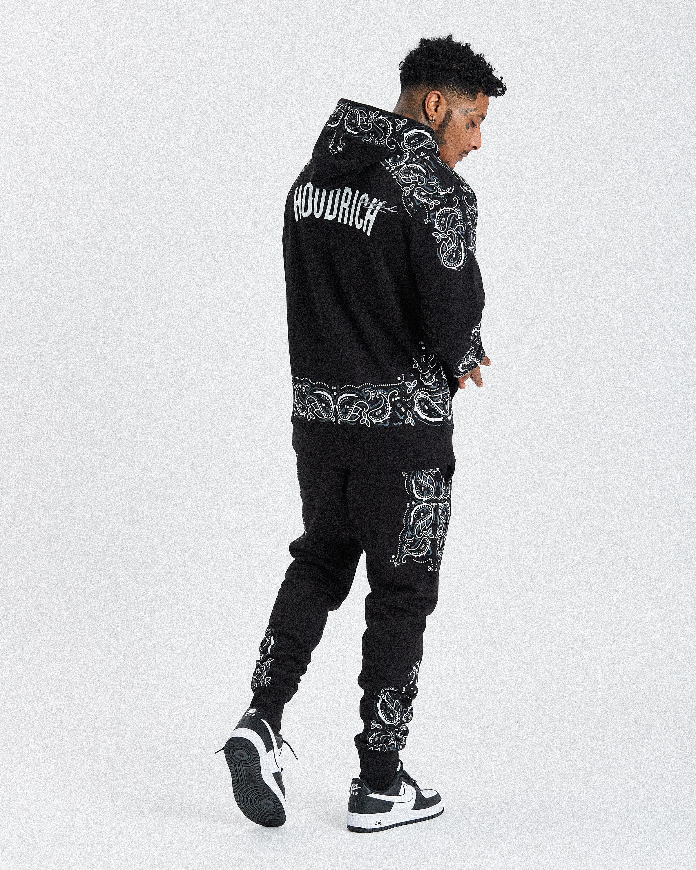 A man wearing a black hooded sweatshirt and matching jogger pants with white ornate graphic prints and the word 'HOODRICH' on the back, shown from a rear three-quarter angle against a light gray studio background.