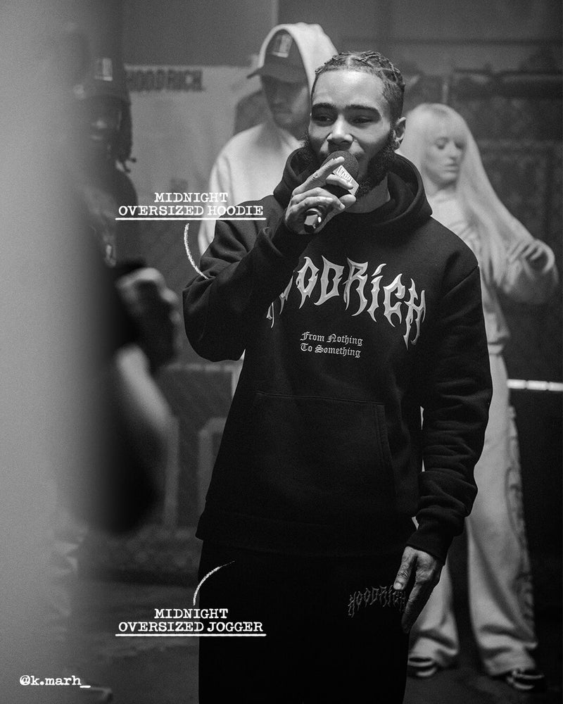 A black and white photo of a man in a hoodie holding a microphone, with other people blurred in the background. The hoodie features the word 'HOODRICH' in a stylized gothic font.
