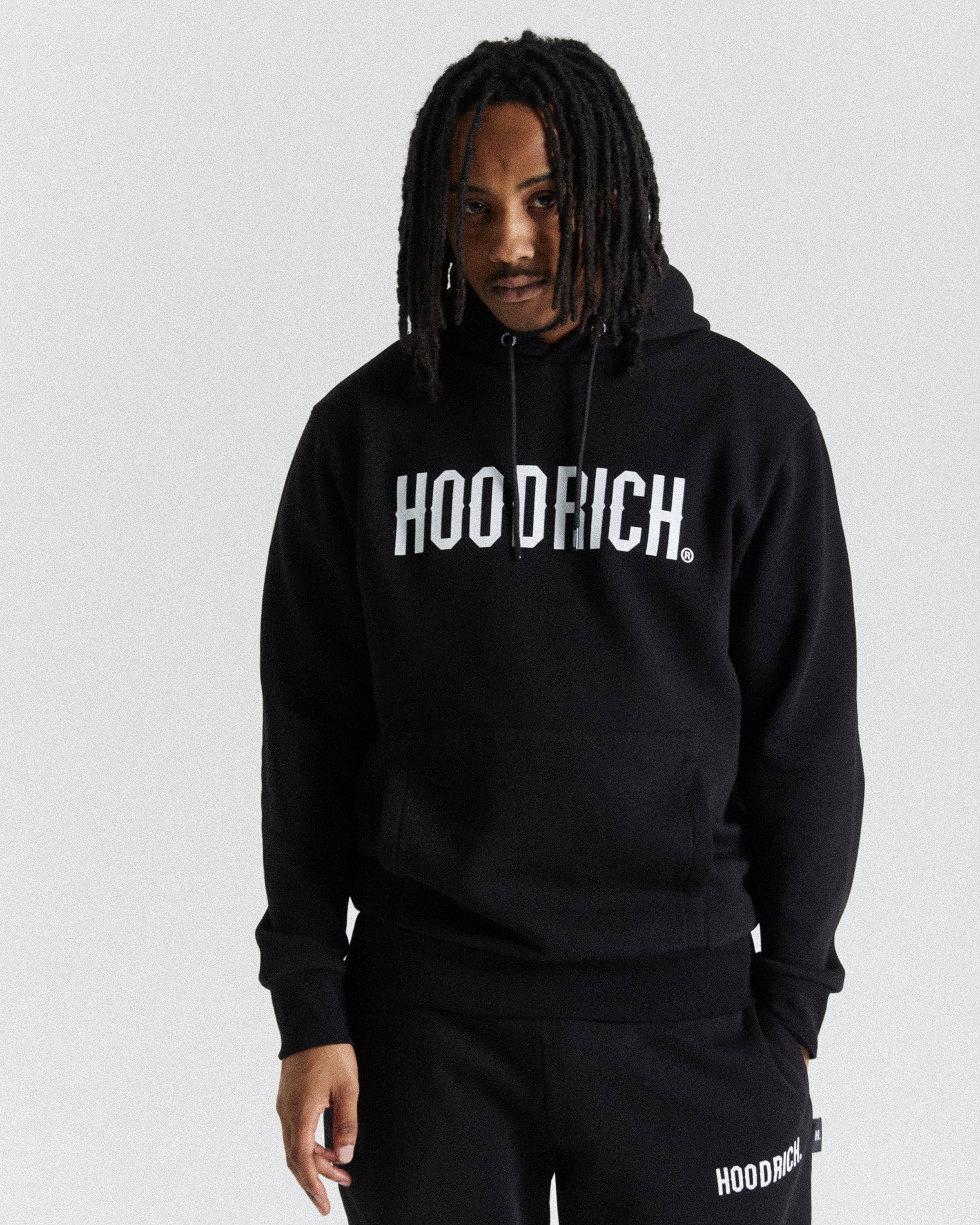 A man wearing a black pullover hoodie with 'HOODRICH.' printed in white across the chest, paired with matching black sweatpants against a light gray studio background.