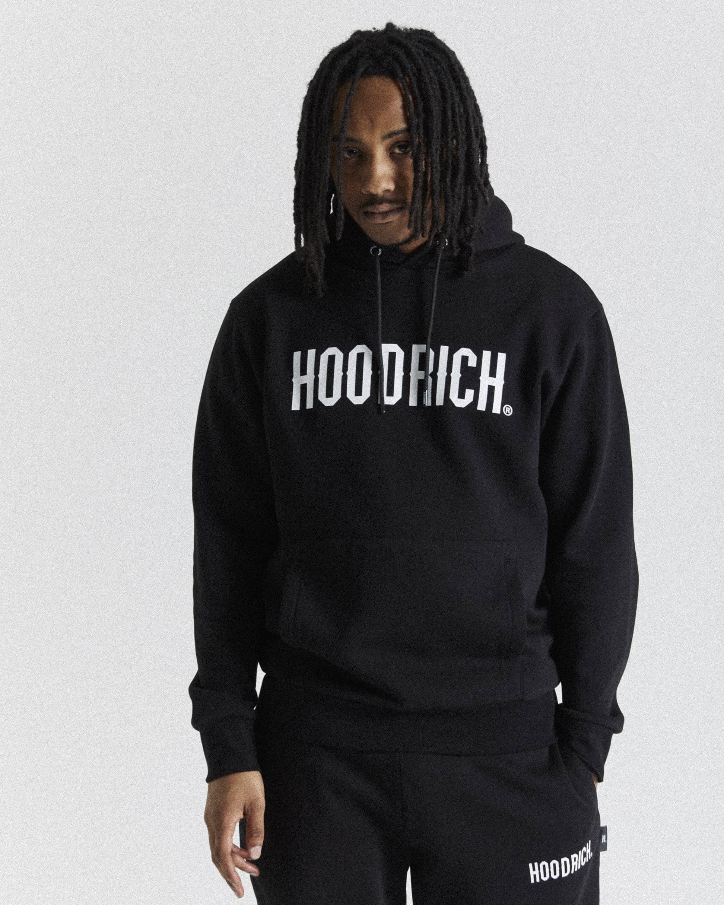 A man wearing a black pullover hoodie with 'HOODRICH.' printed in white across the chest, paired with matching black sweatpants against a light gray studio background.