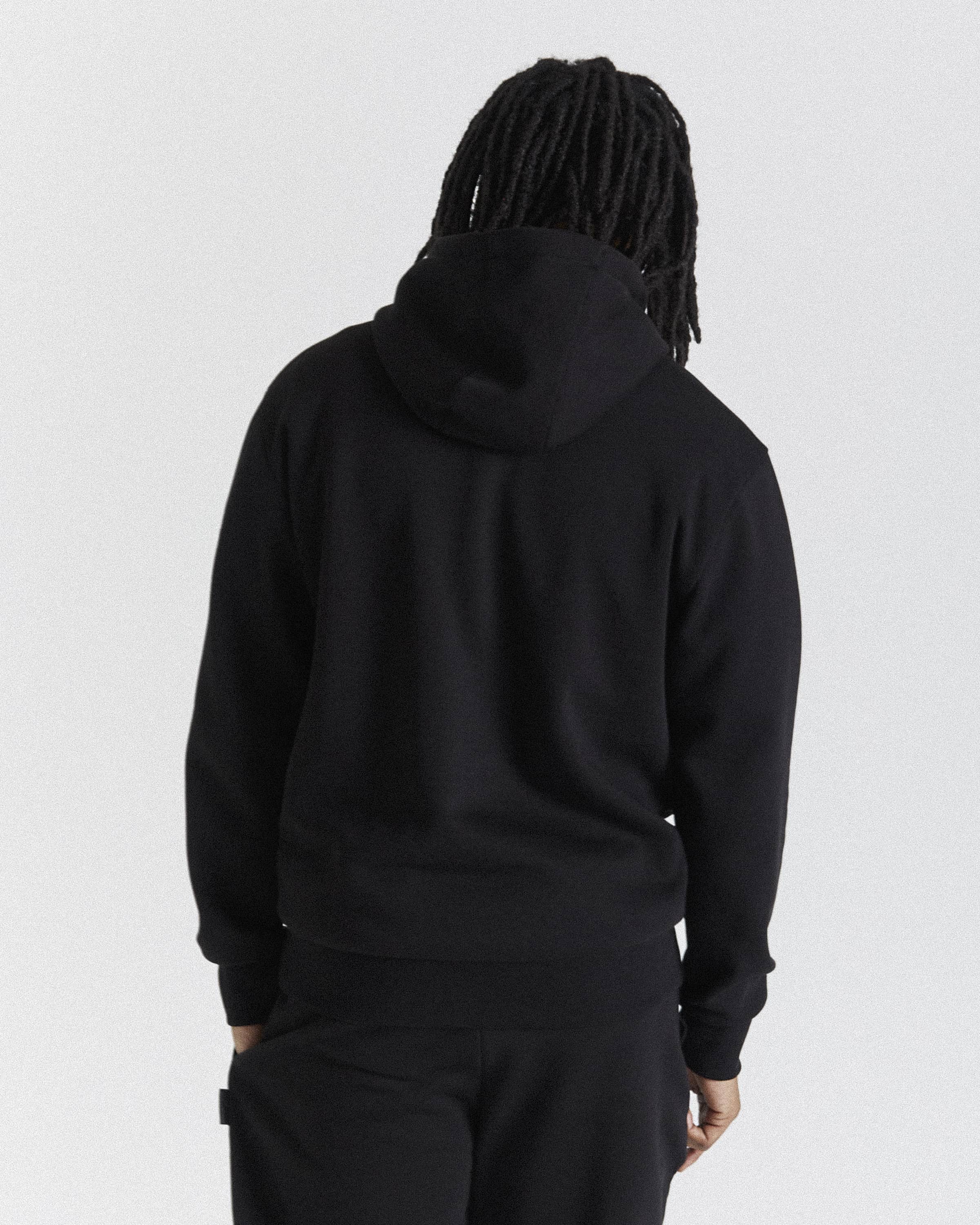 A person viewed from the back wearing a black pullover hoodie and black pants against a plain light gray studio background.