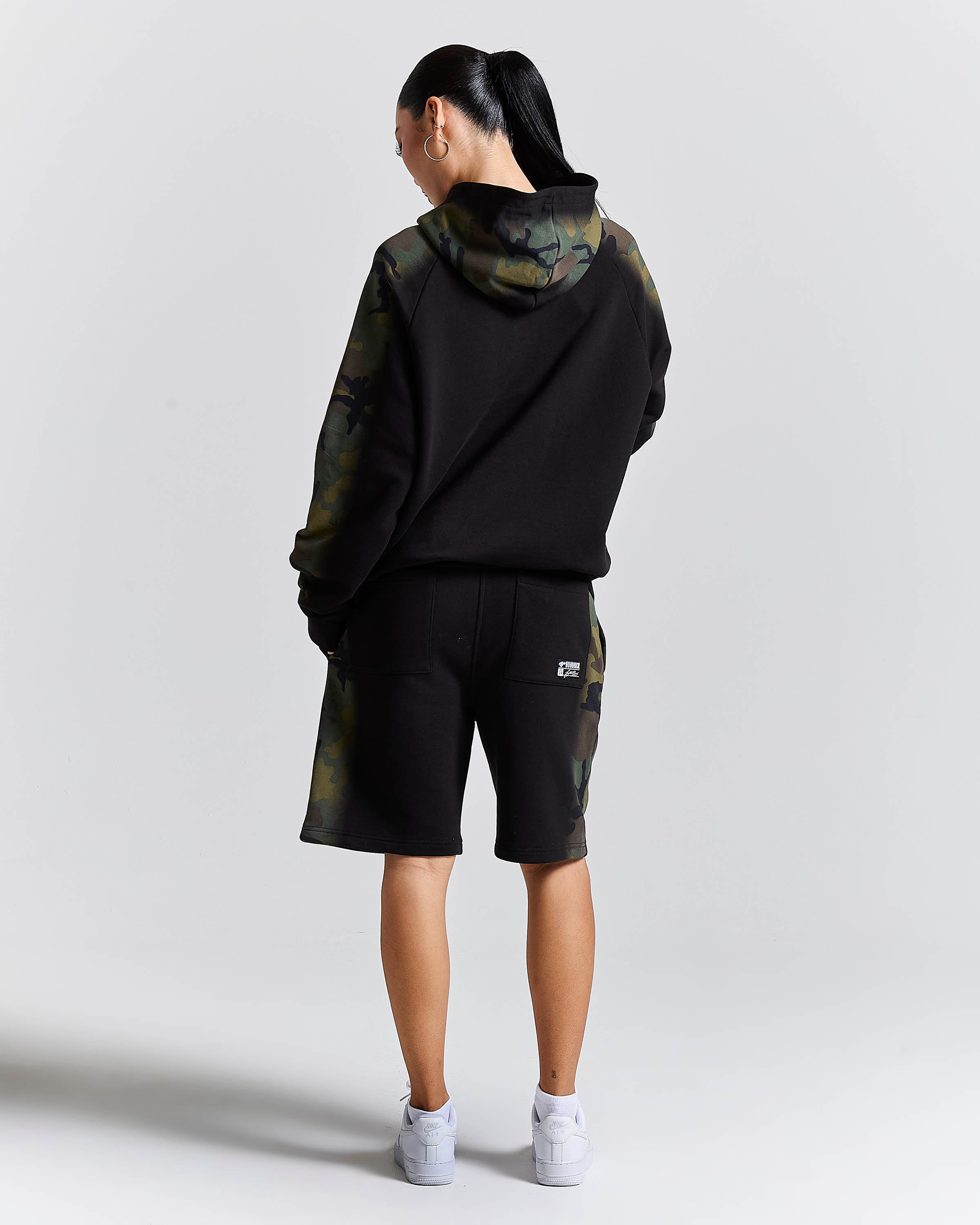 A model shown from the back wearing a black hoodie with olive green camouflage sleeves and hood, matching camouflage shorts, and white sneakers, set against a plain light gray studio background.