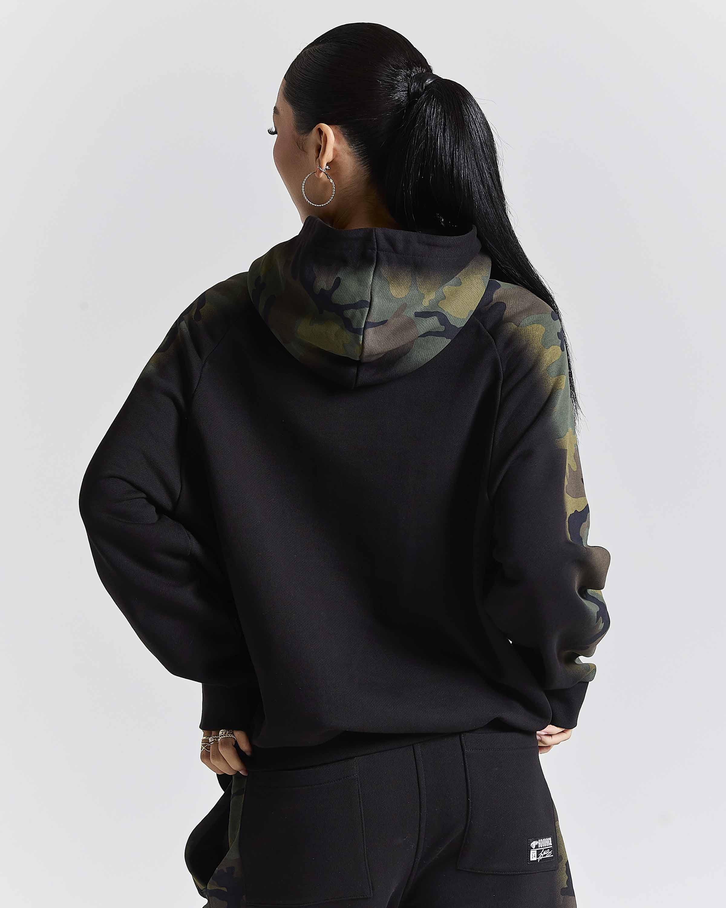 A woman shown from the back wearing a black hoodie with a green and brown camouflage pattern on the hood and upper sleeves, paired with black sweatpants against a light gray studio background.
