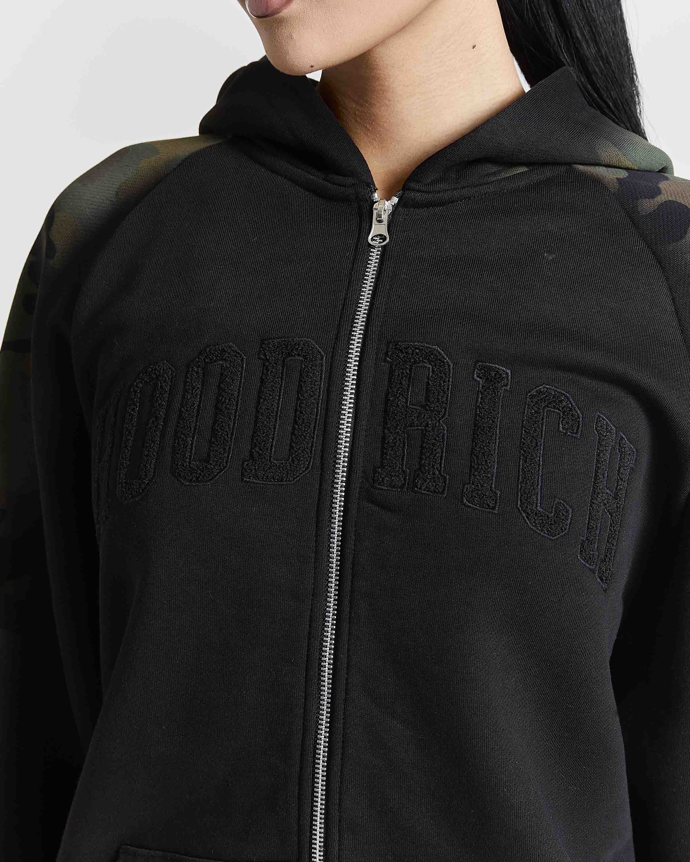A close-up of a person wearing a black zip-up hoodie with 'HOOD RICH' embroidered in arched black letters across the chest. The hoodie features a silver zipper, a black hood, and subtle camouflage pattern details on the shoulders.