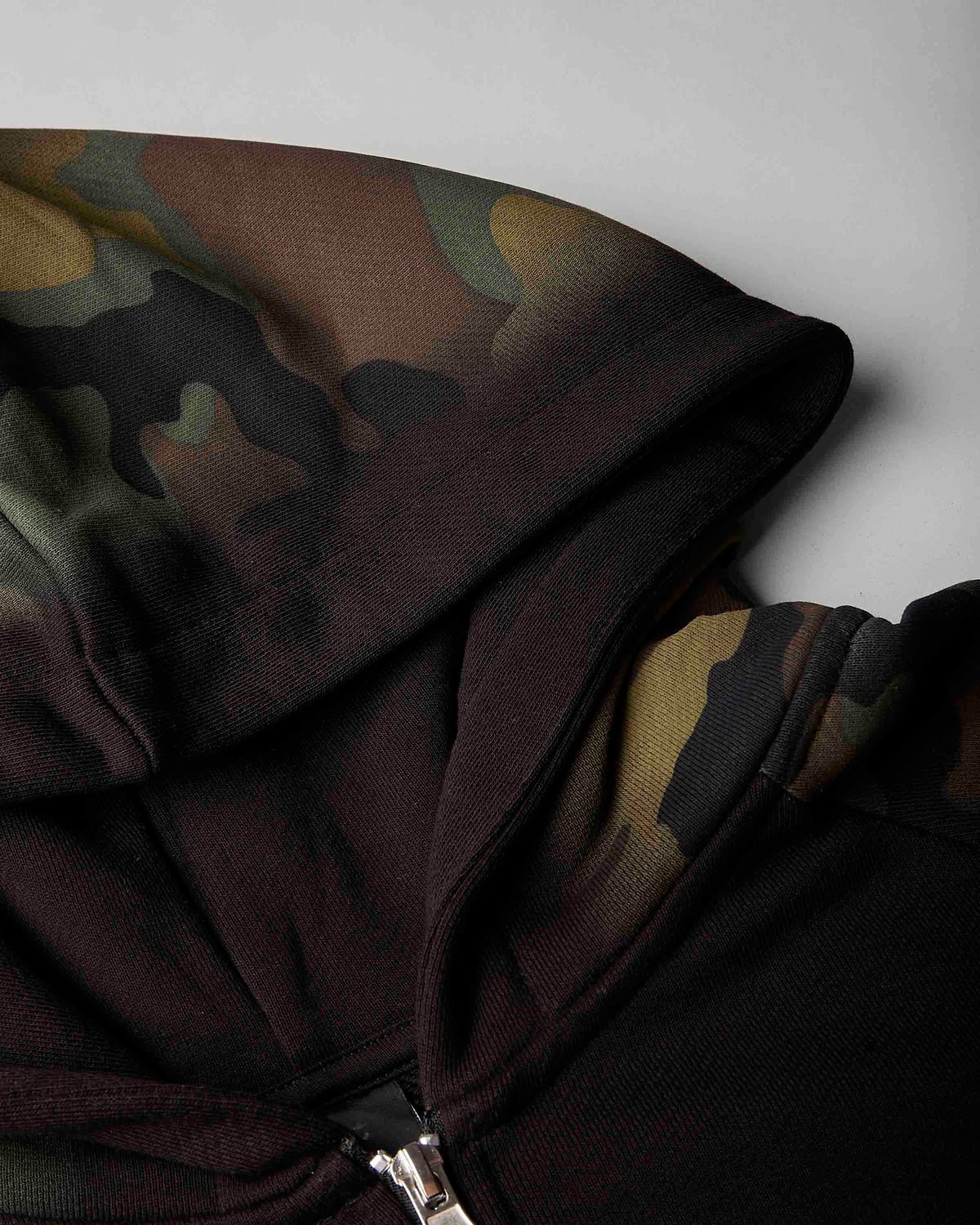Close-up of a hooded sweatshirt showing a dark camouflage pattern on the hood and shoulder, with a black lower body and a silver zipper.