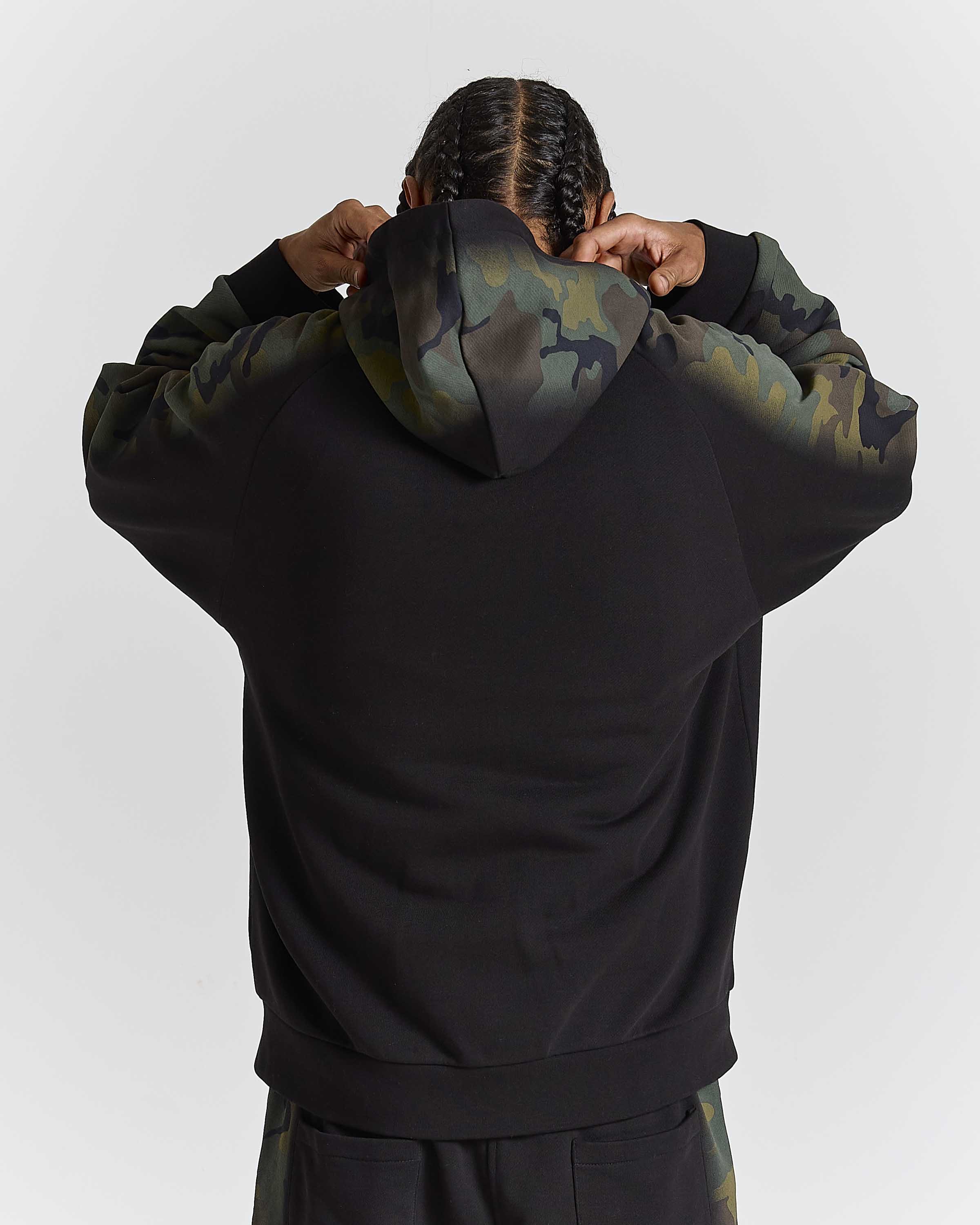 A person viewed from behind wearing a black hoodie with camouflage-patterned hood and sleeves, standing against a light gray studio background.
