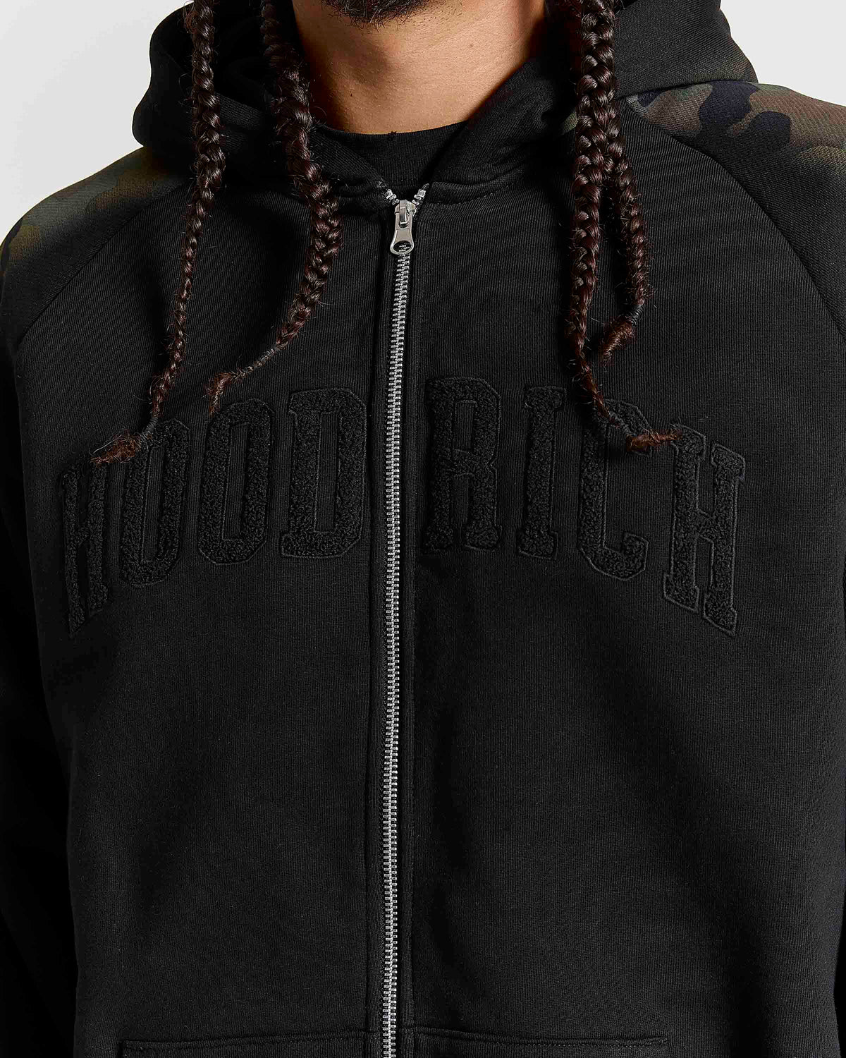 A close-up of a person wearing a black zip-up hoodie with 'HOODRICH' embroidered in tonal black across the chest. The hoodie has a silver zipper, black drawstrings, and a subtle camouflage pattern on the right shoulder.