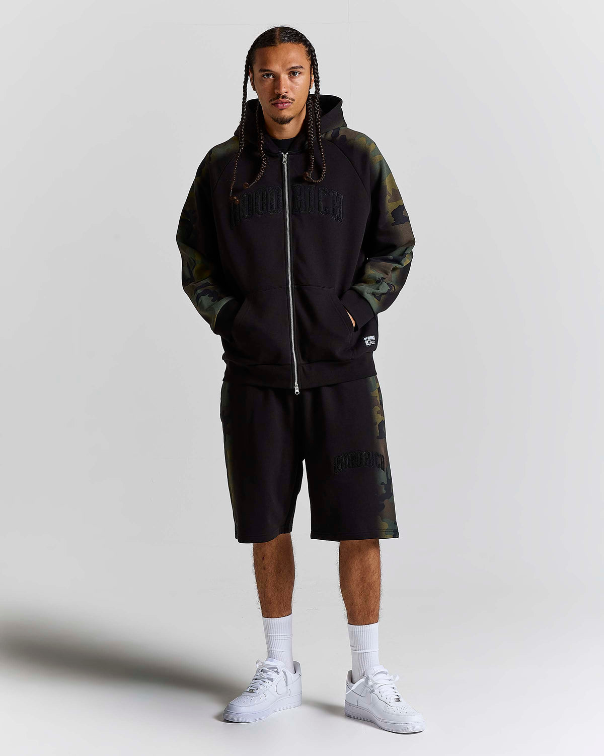 A man modeling a black zip-up hoodie and matching shorts set from HOODRICH, featuring dark green camouflage sleeves and side panels, styled with white sneakers and socks against a light gray studio background.