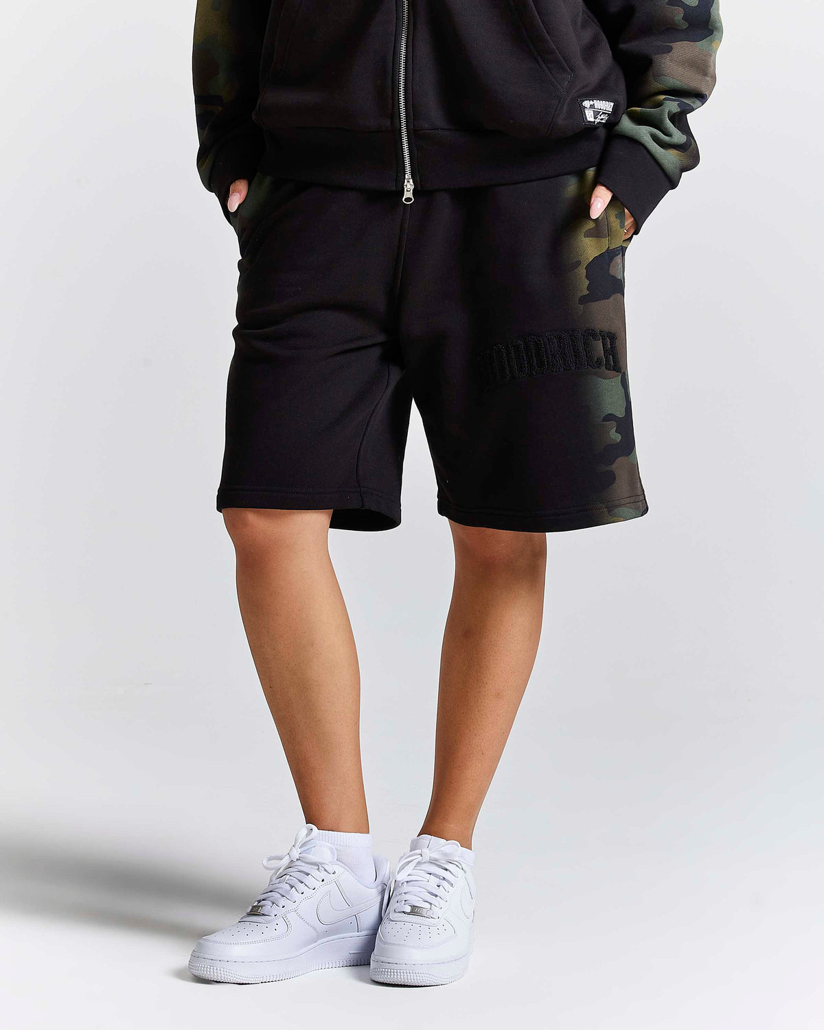 A person wearing black sweat shorts with a green camouflage side panel and 'HOODRICH' text, paired with a black zip-up hoodie and white sneakers, against a light gray studio background.