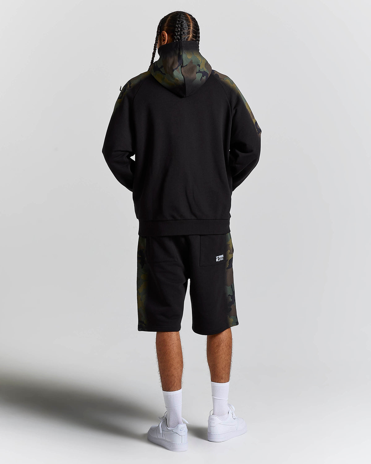 A model shown from the back wearing a black hoodie with camouflage pattern details on the hood and sleeves, matching camouflage shorts, white socks, and white sneakers, set against a plain light gray studio background.