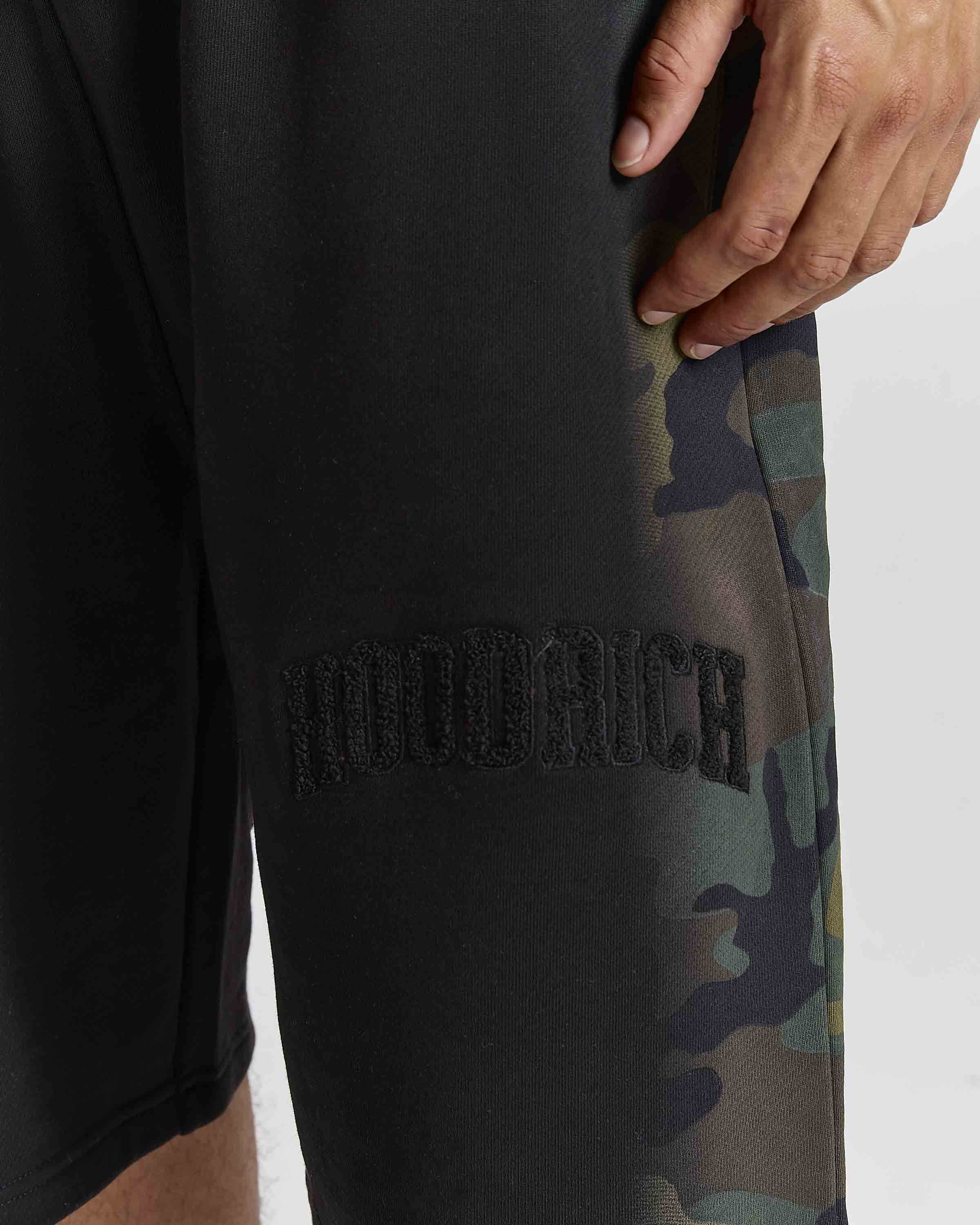 Close-up of a person wearing black sweatpants with a tonal black embroidered 'hoodrich' logo and a green camouflage pattern along the side, against a light gray background.