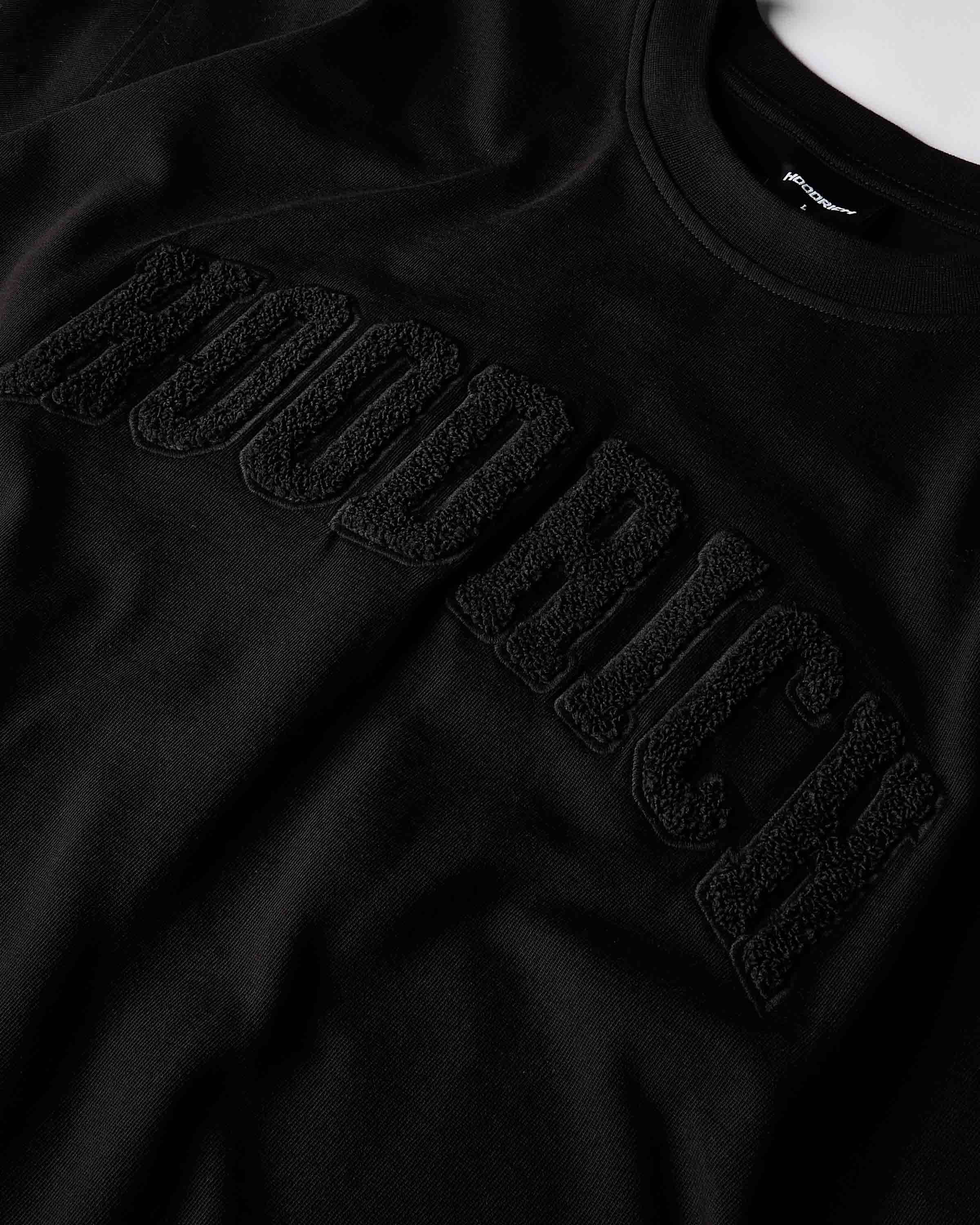 Close-up of a black crewneck sweatshirt featuring a large, tonal black embroidered 'HOODRICH' logo across the chest