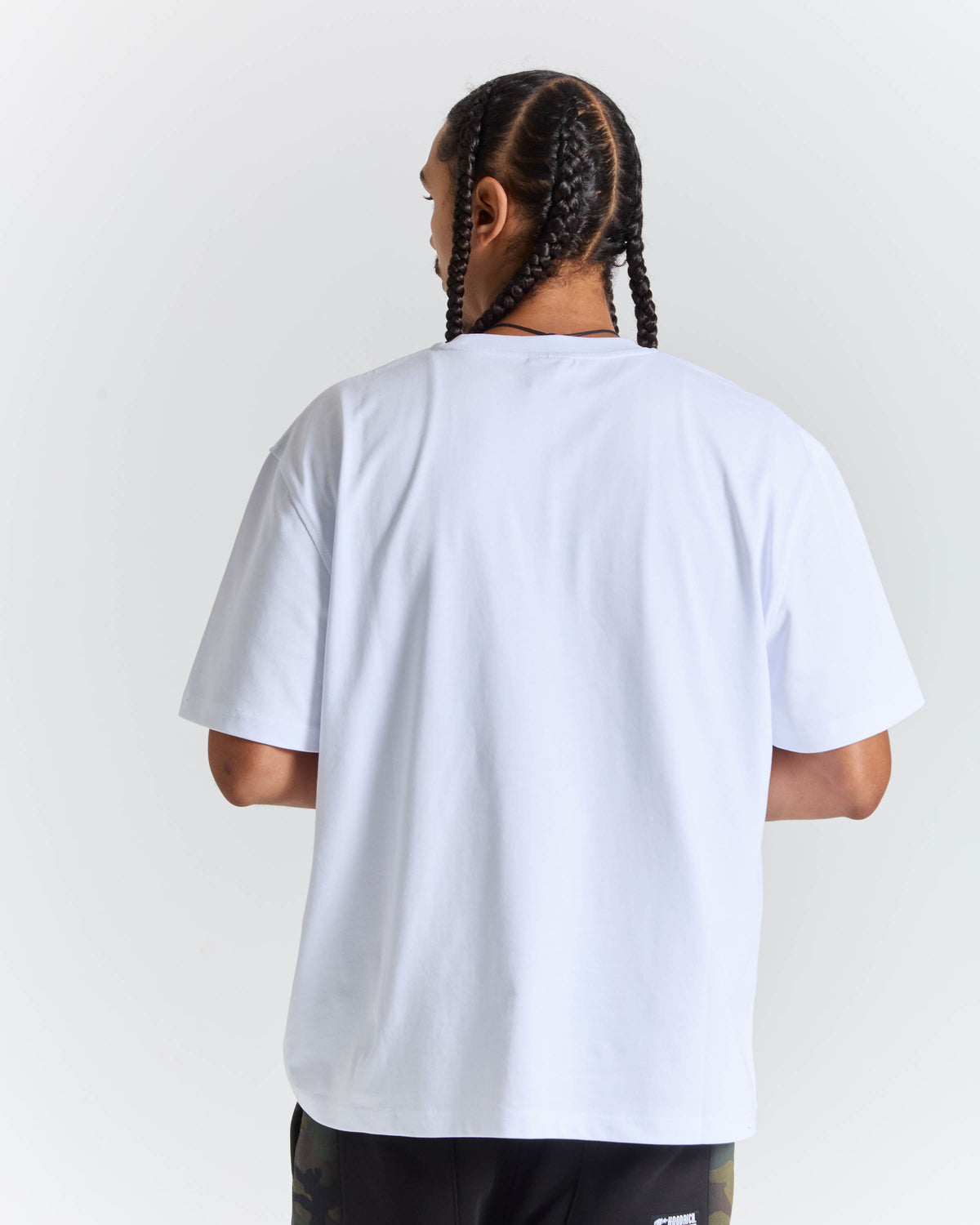 A man with braided hair is shown from the back wearing an oversized white t-shirt and dark patterned shorts against a light gray studio background.
