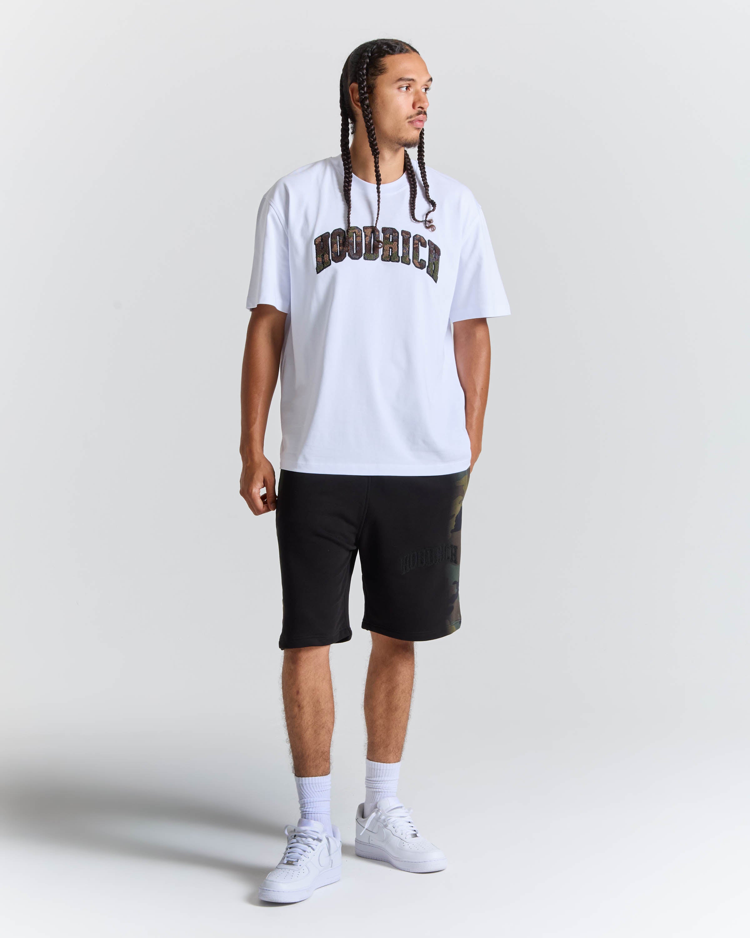 A man modeling a white hooded t-shirt with 'HOODRICH' printed in dark brown across the chest, paired with black shorts, white socks, and white sneakers, set against a plain light gray studio background.