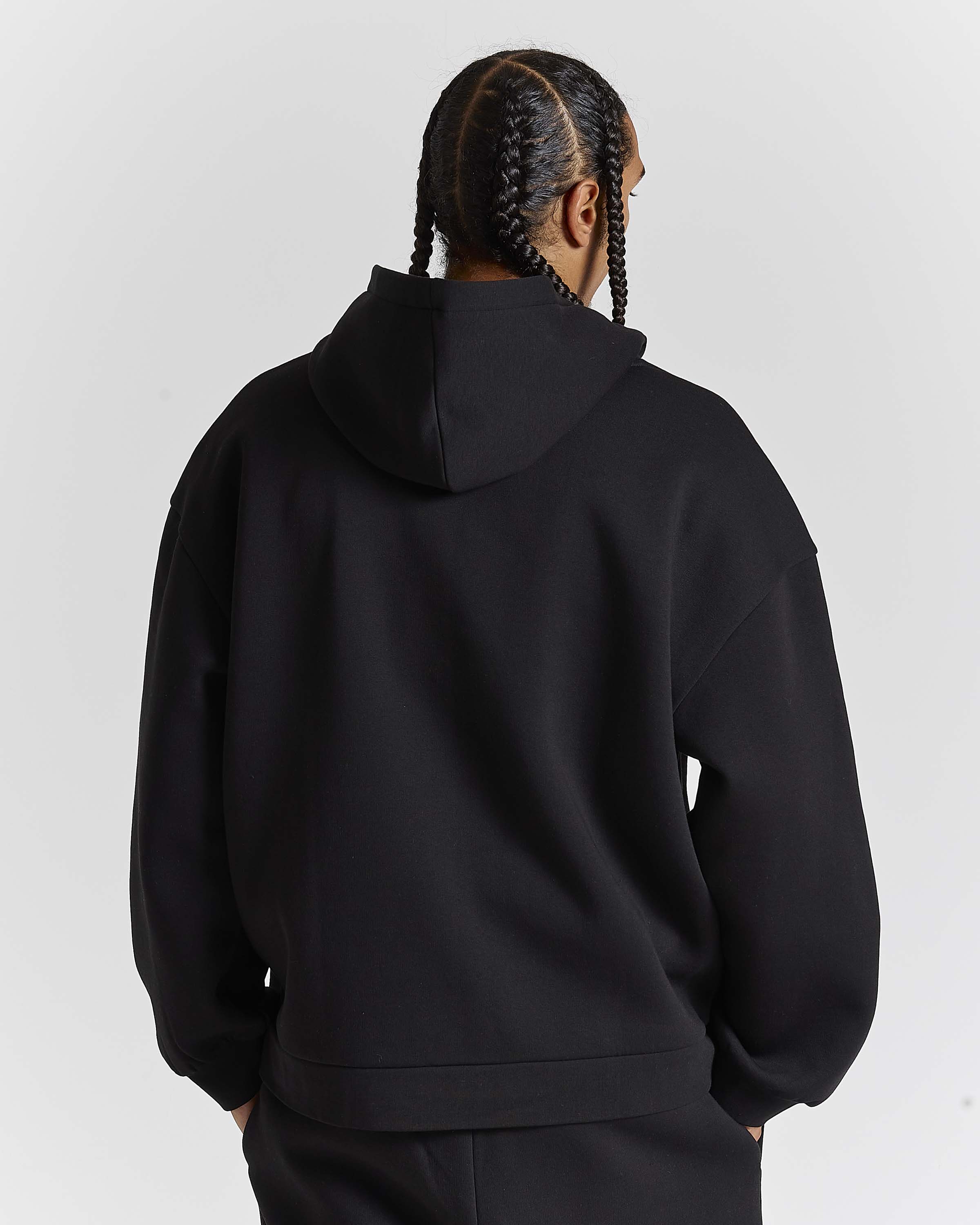 A person shown from the back wearing a black pullover hoodie and matching black pants against a light gray studio background.