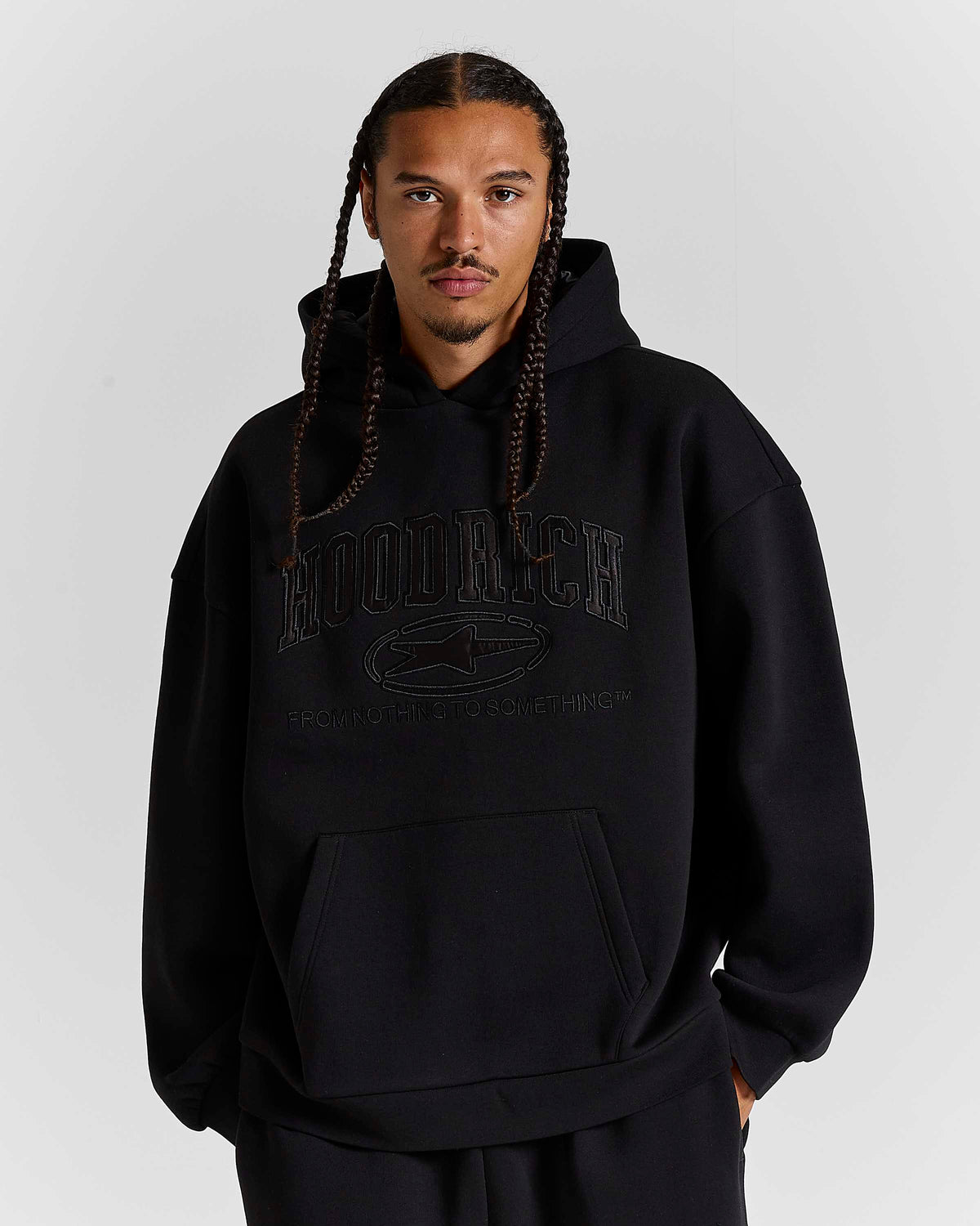 A man with braided hair wearing a black hooded sweatshirt with tonal black graphic text reading 'HOODRICH' and a logo on the chest, photographed against a light gray studio background.