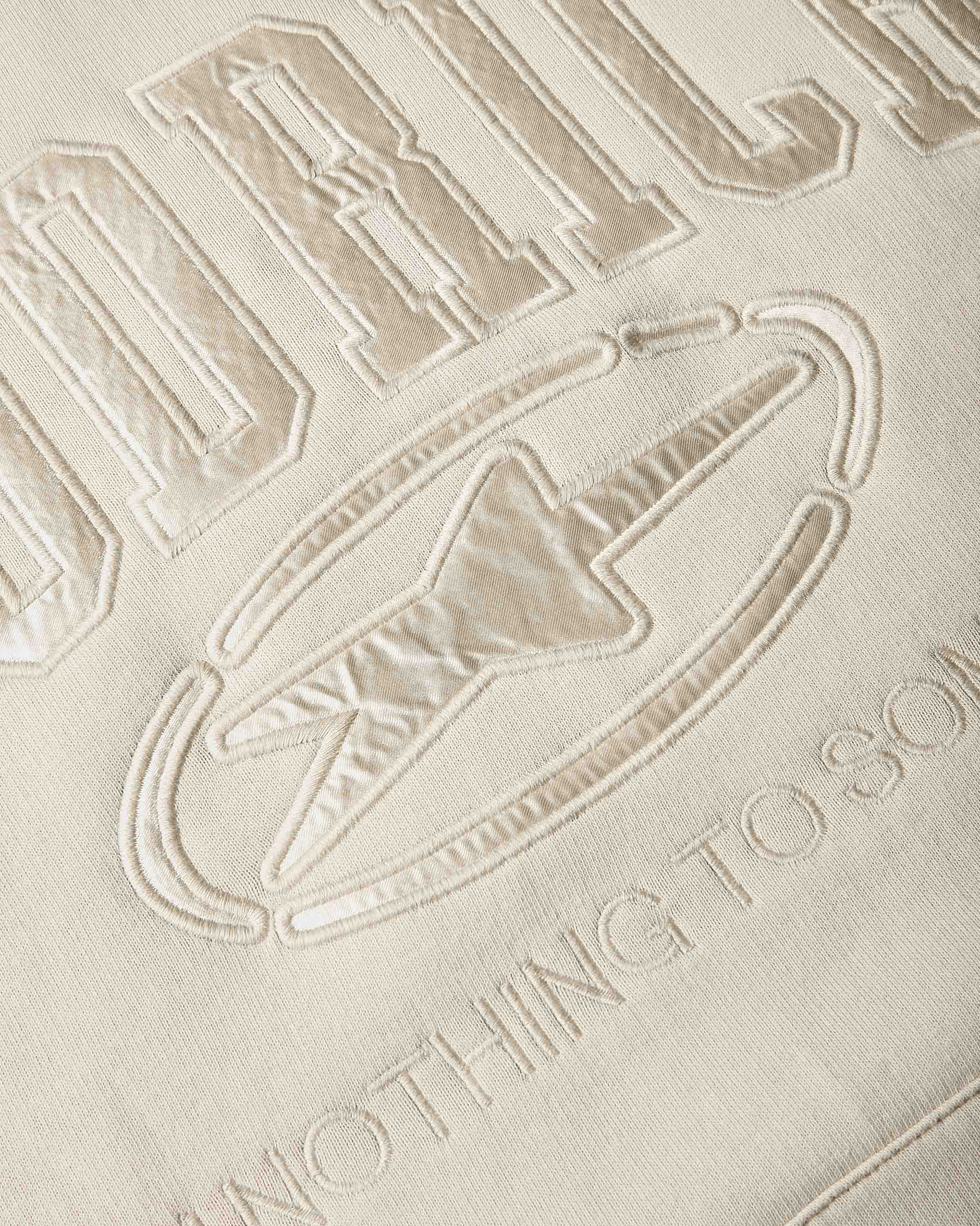 Close-up of a beige embroidered fabric with a large stylized letters and a logo outlined in raised stitching.