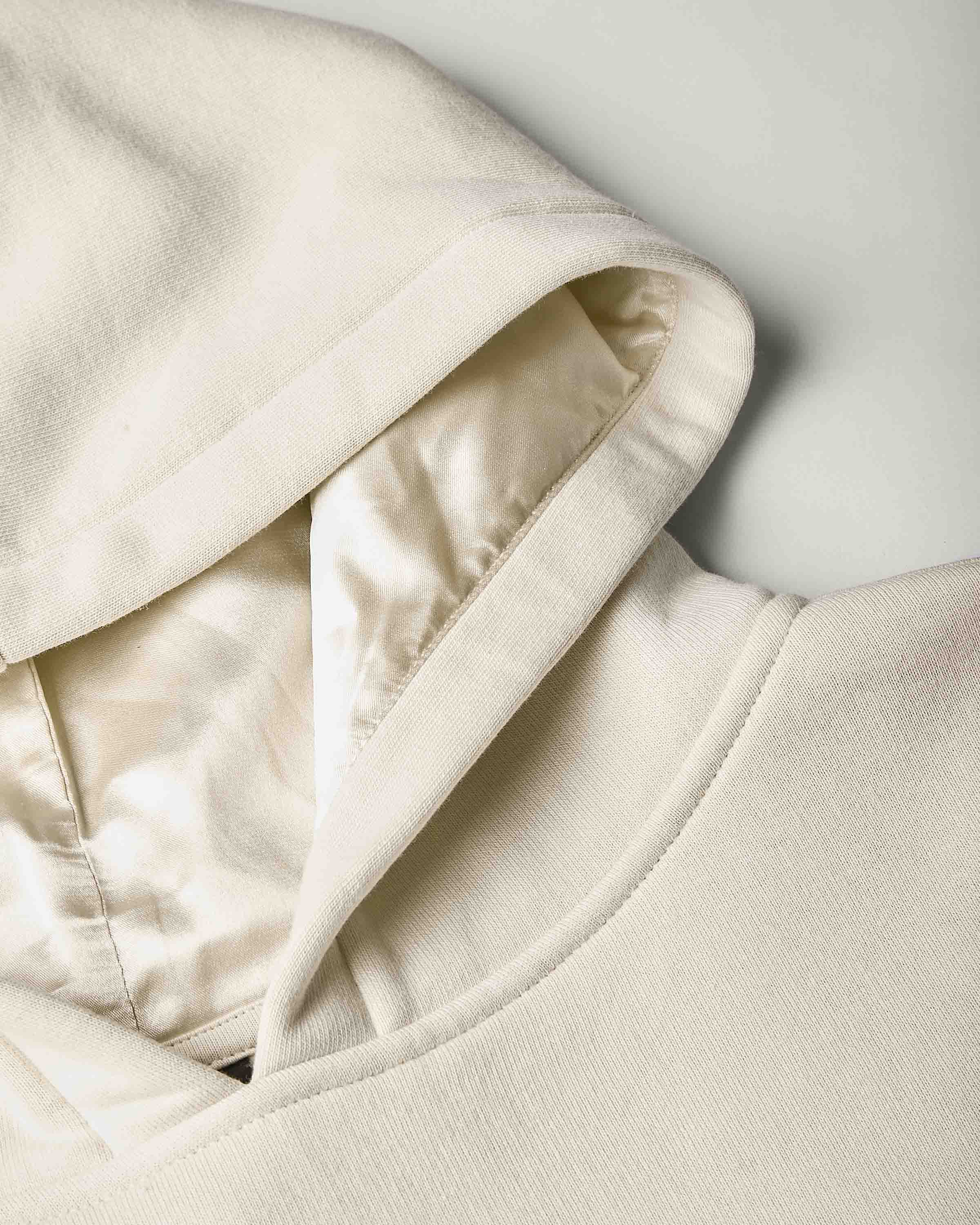 Close-up of a cream-colored hooded sweatshirt showing the hood satin lining, ribbed knit fabric, and soft interior details against a light gray background.