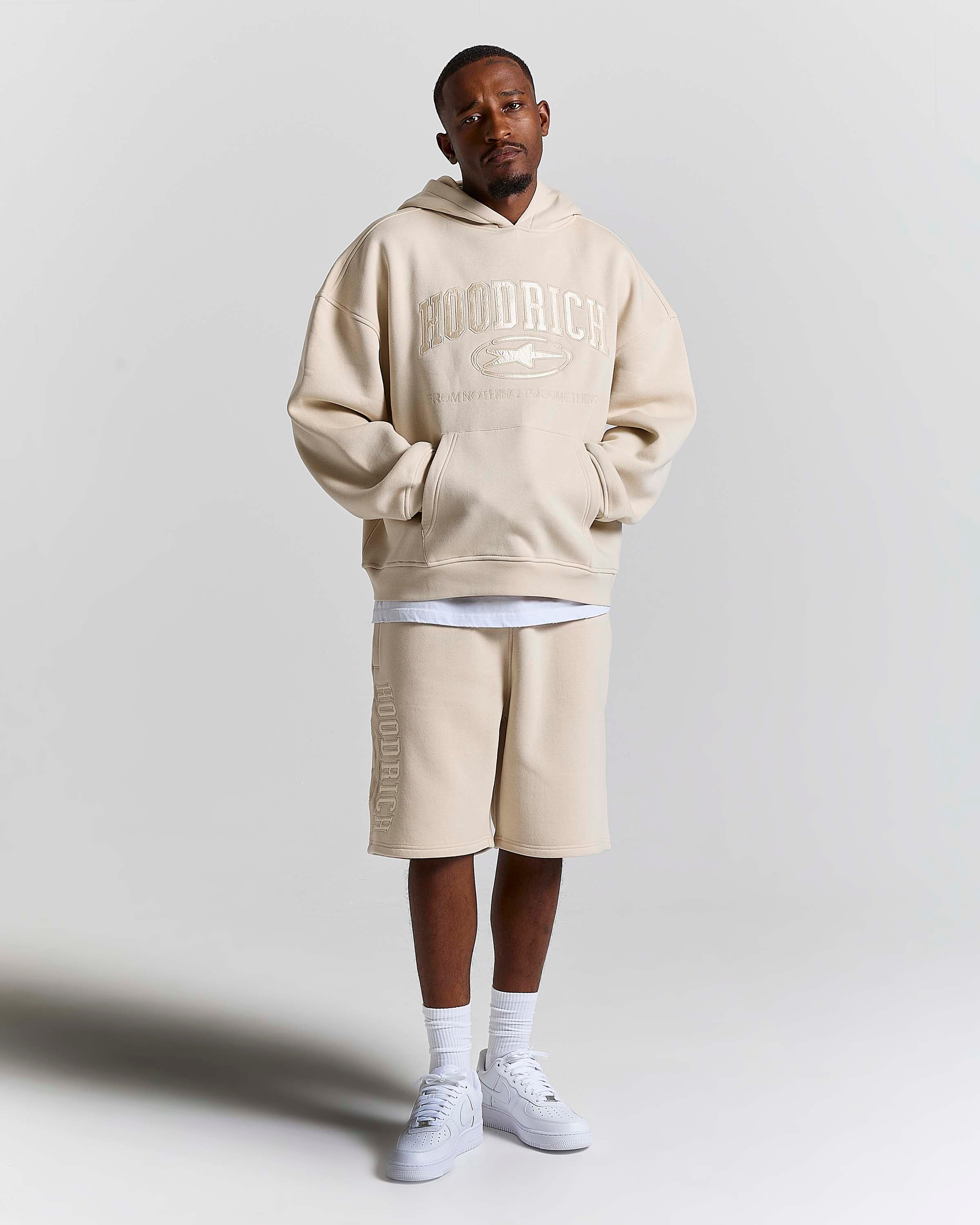 A man modeling an oversized beige hoodie with 'HOODRICH' text and a matching beige drawstring shorts set, styled with white t-shirt layers, white socks, and white sneakers against a light gray studio background.