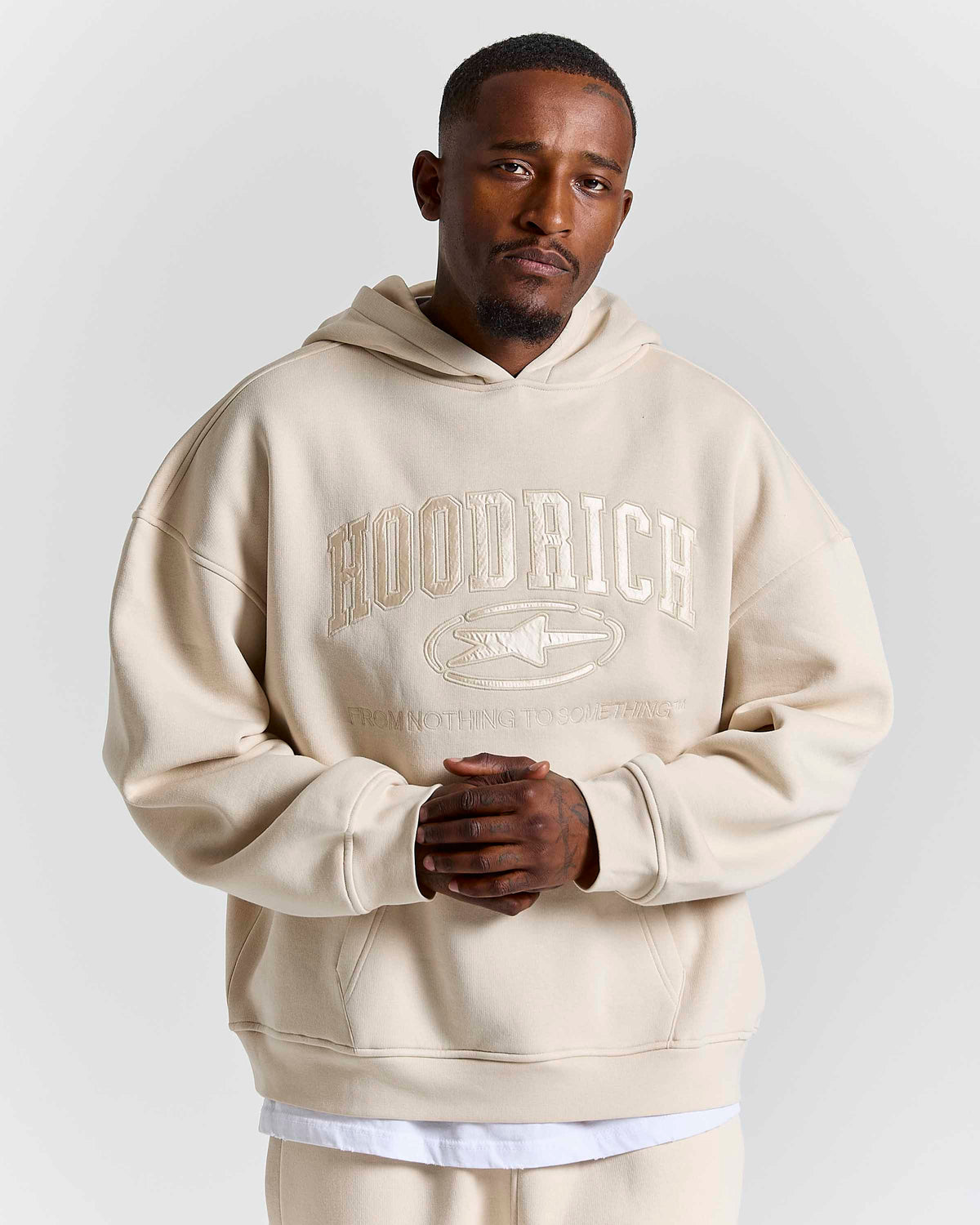 A man wearing an oversized beige hoodie with a tonal 'HOODRICH' logo and fish graphic on the chest, paired with matching beige pants, against a light gray studio background.