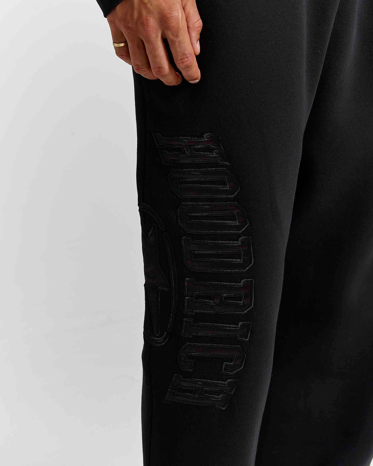 Close-up of a person wearing black sweatpants with large tonal black 'hoodrich' lettering on the side, shown against a light gray studio background.