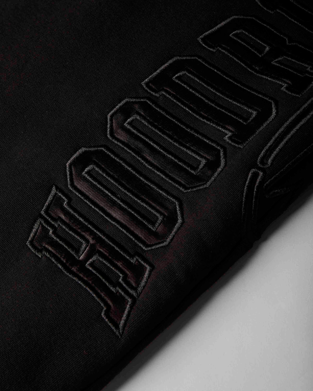 Close-up of a black fabric surface with large tonal embroidered lettering running diagonally across the frame.