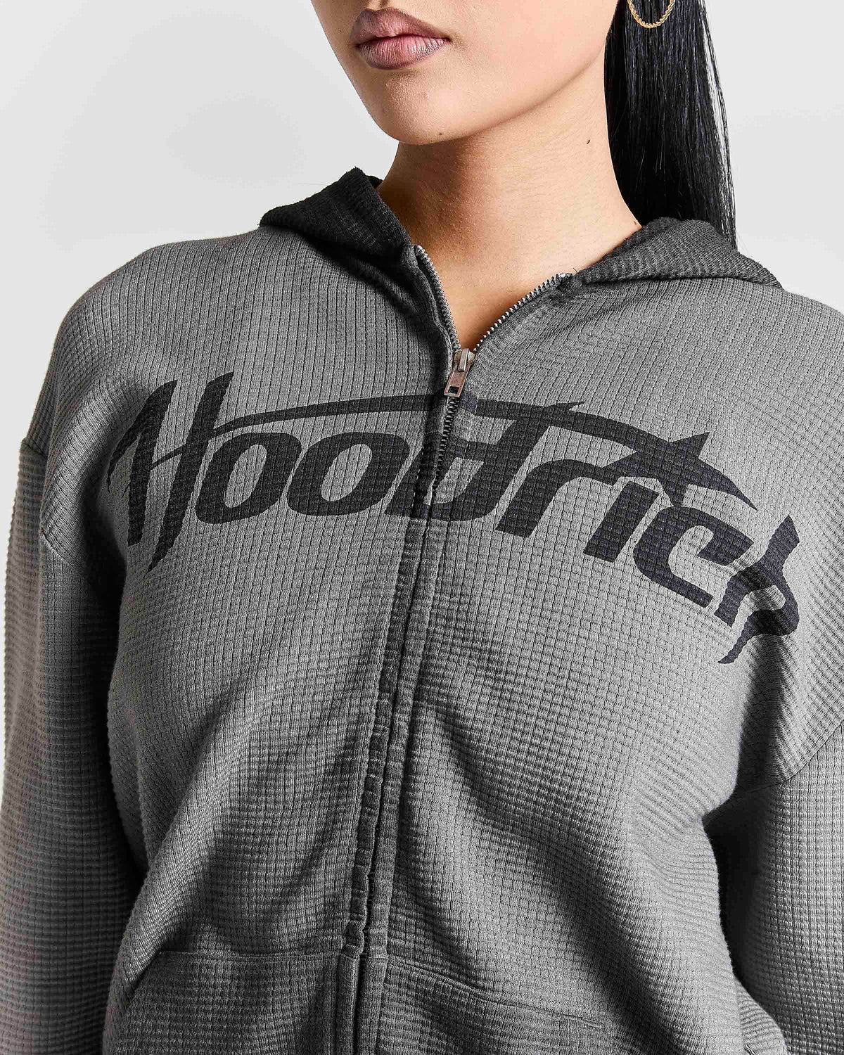 A close-up of a person wearing a gray waffle-knit zip-up hoodie with a large black 'Hoodrich' logo across the chest, shown against a plain light background.